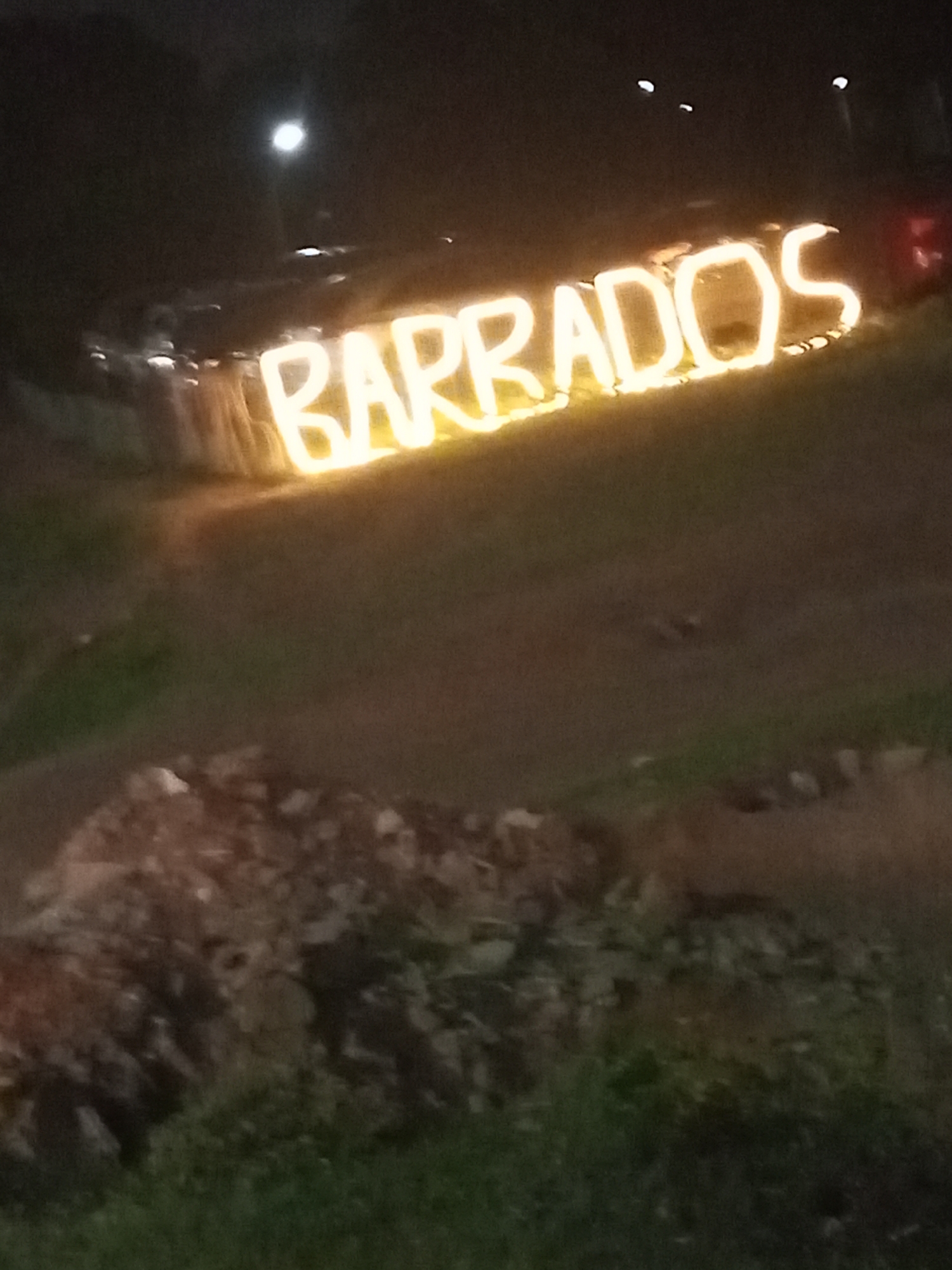 The image shows the illuminated word "BARRADOS" at night, likely in Abuja, Nigeria. The letters are bright and seem to be made of light tubes or similar material. In the background, there are blurred streetlights and possibly vehicles. The foreground contains a dark patch of ground with what appears to be some kind of vegetation or ground cover. The overall scene suggests a place of business or a location advertising its name at night.