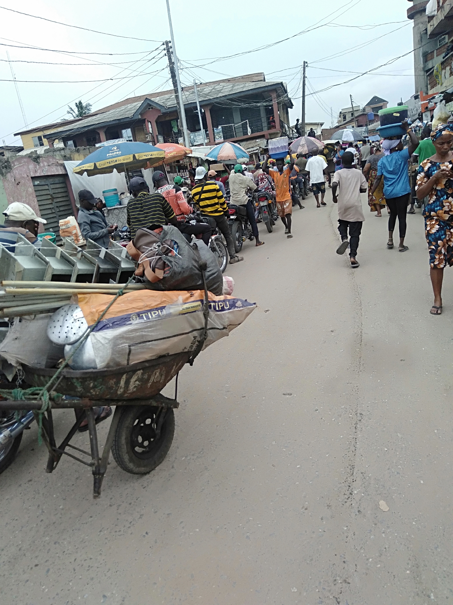 The image captures a bustling street scene in Shangisha, Nigeria. The foreground is dominated by a heavily laden, rusted wheelbarrow filled with an assortment of items, including metal cookware, bamboo poles, and sacks with the inscription "TIPU". The wheelbarrow is positioned on the left side of the frame, partially obscuring a parked motorcycle.
The middle ground reveals a dense throng of people and motorcycles filling the street. A row of motorcycles with riders wearing casual clothing and helmets are visible behind the wheelbarrow. Several individuals are walking along the street, some carrying items on their heads. Market umbrellas provide shade for roadside vendors and their customers.
The background features a mix of low-rise buildings with corrugated metal roofs and overhead power lines that crisscross the sky. The environment suggests a typical urban marketplace setting, filled with activity and commerce.