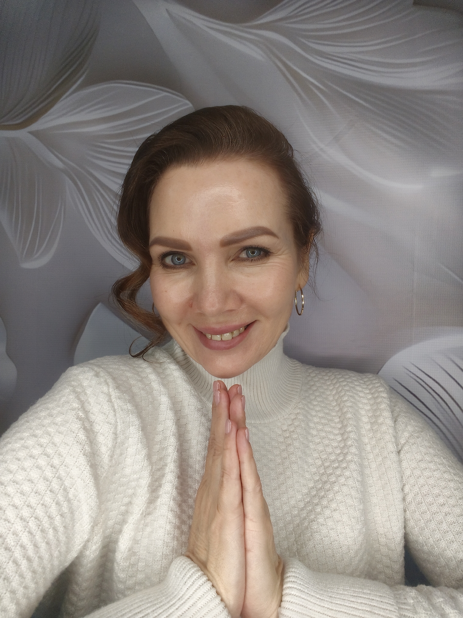A woman with brown hair and blue eyes is smiling at the camera with her hands pressed together in a gesture of prayer or greeting. She is wearing a white turtleneck sweater and hoop earrings. The background is a light gray and white floral pattern. The location is unknown.