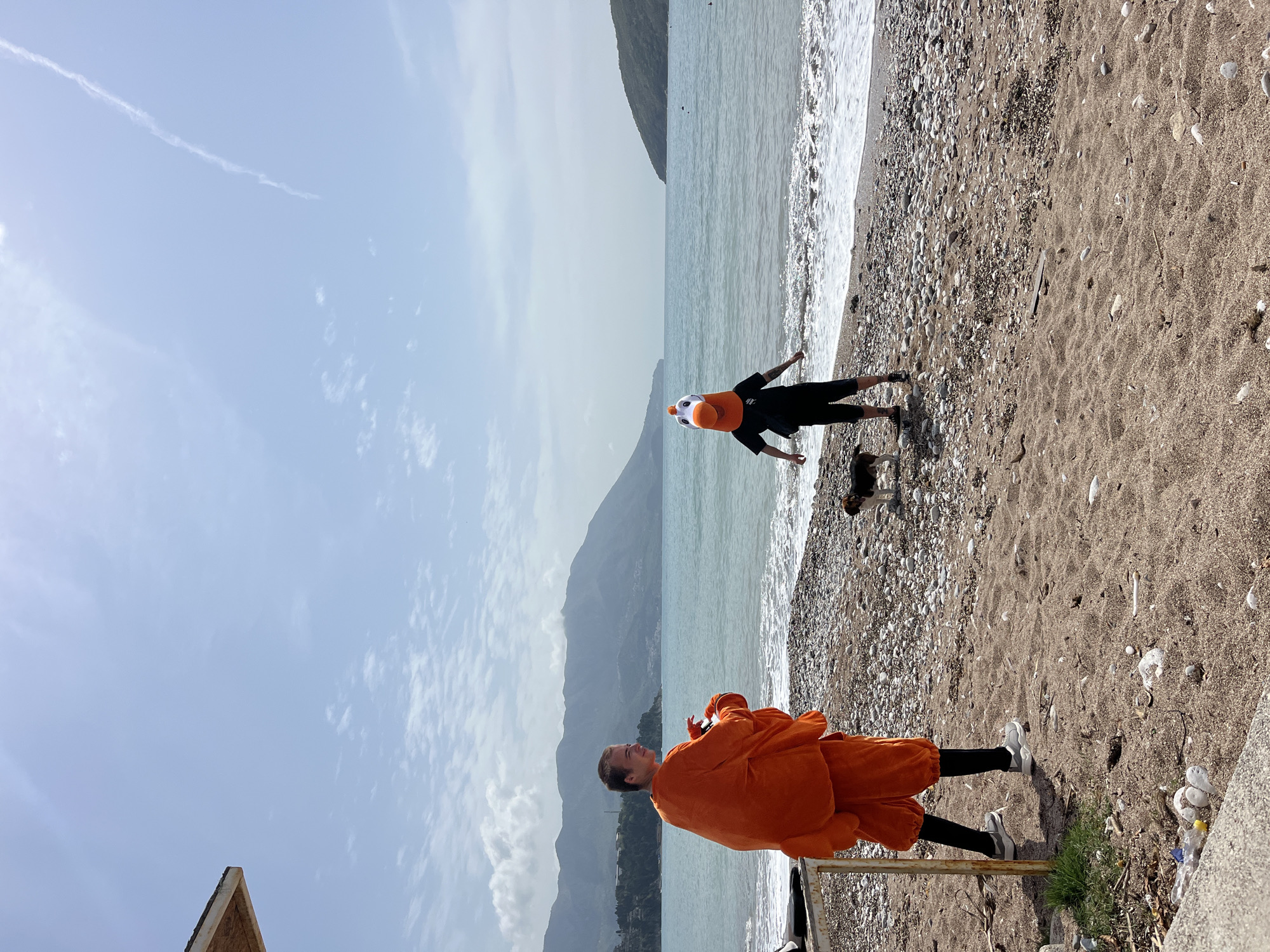 The image depicts a scene on a beach with a mountainous backdrop. A person in an orange chicken costume stands near the right side of the frame, while another person in black clothing and a duck hat stands near the water's edge further away. A small dog is also present near the water. The beach consists of sand and pebbles. The sky is partially cloudy, and a mountain range is visible in the background. The location is in the city of Budva, Montenegro.