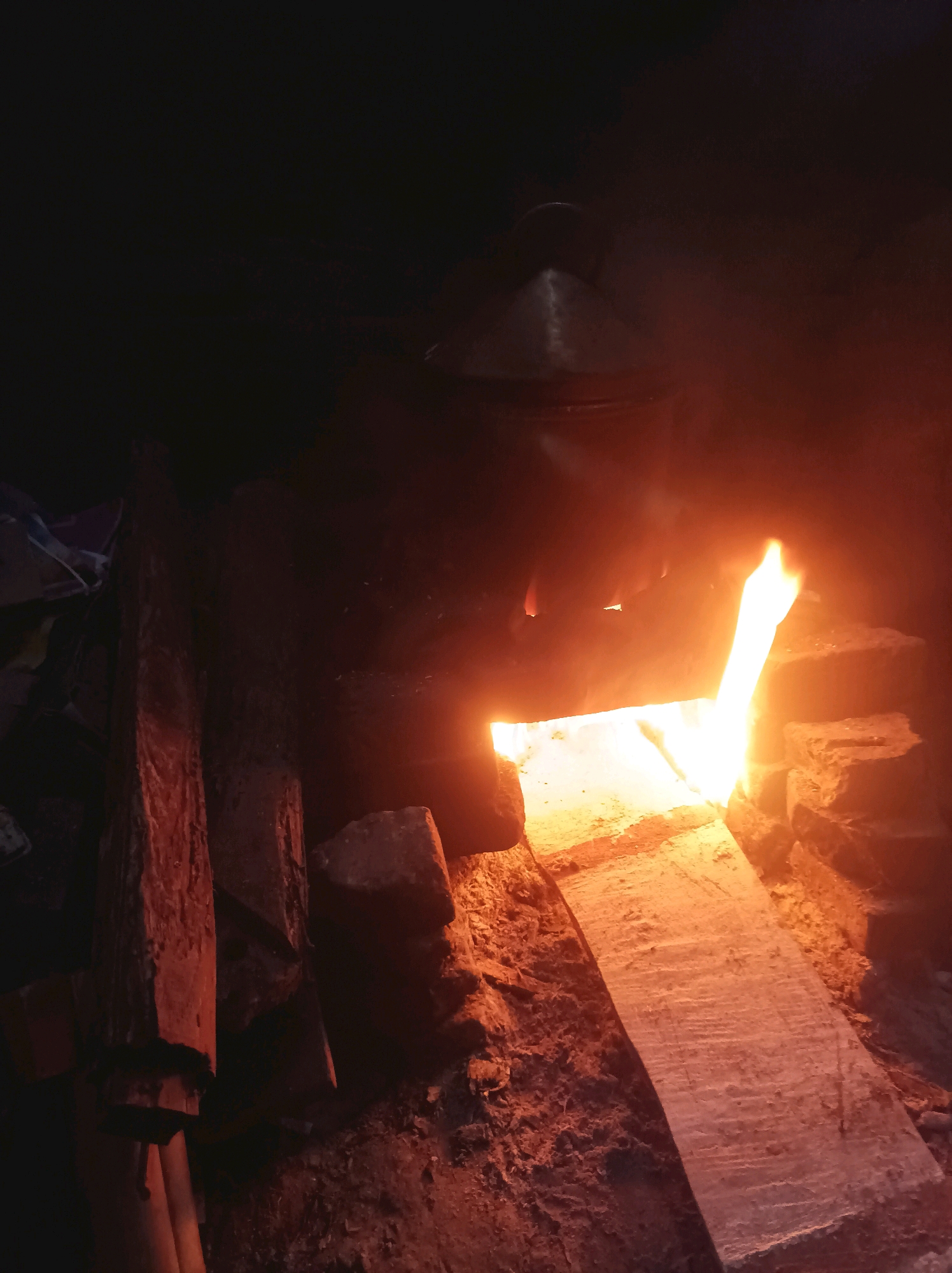 An image captures a close-up view of a rustic, brick-built oven with a fire burning inside, casting a warm, orange glow. A dark, possibly metal, pot or kettle rests on top of the oven. The environment appears to be outdoors, with natural materials like rough-hewn wood and possibly dirt visible in the foreground and surrounding the oven. The scene is dimly lit, with the fire being the primary light source, creating shadows and highlighting the textures of the brick and wood. The overall impression is one of a functional, perhaps traditional, cooking setup in a natural or less developed setting. The location within Canga'an, Indonesia, suggests a potentially rural or traditional context for this type of oven.