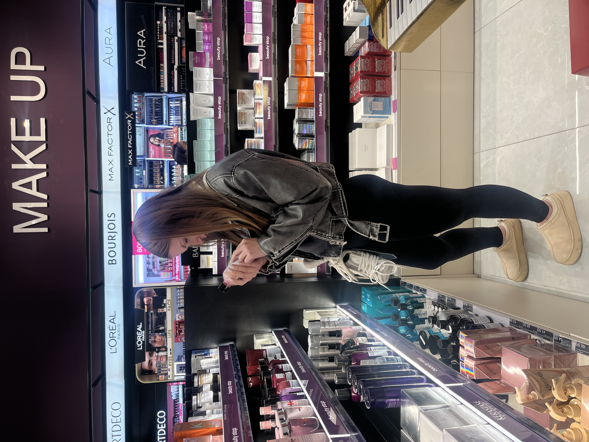 The image depicts a young adult female examining a cosmetic product within a brightly lit beauty retail store in Budva, Montenegro. The female subject, positioned slightly right of center, has long brown hair and is wearing a grey distressed denim jacket, black leggings, and beige slip-on shoes with thick soles. She holds a light-colored bottle with a dark cap in both hands, orienting it downwards as she inspects its label or contents. A white, coiled object, possibly a strap or shoelaces, hangs from her left shoulder.
The store's environment features extensive display shelving showcasing numerous beauty brands. A large, illuminated sign overhead prominently displays "MAKE UP." Below this, horizontal illuminated panels list brand names including "ARTDECO," "L'ORÉAL PARIS," "BOURJOIS," "MAX FACTOR X," and "AURA." Products from these brands, as well as "Kérastase," "YOSKINE," "BABOR," and "CENTELLA," are neatly arranged on multiple tiered shelves equipped with LED strip lighting. White labels marked "beauty stop" and various price indicators are visible on the shelf edges. The floor is covered with large, light-colored, possibly white or light grey, tiled squares. In the lower right foreground, a segment of a decorative golden structure with black and white keys, resembling a stylized piano, is partially visible. The scene suggests a commercial transaction or product selection in progress.
