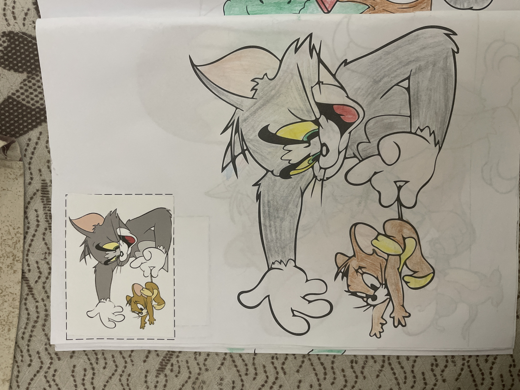 A close-up photograph captures a hand-drawn and colored illustration of Tom and Jerry on white paper, placed over a patterned fabric surface. The main subject is a large depiction of Tom the cat holding Jerry the mouse by his tail. Tom is rendered in shades of grey with black outlines, yellow eyes, and red in his mouth, leaning forward with a distressed or surprised expression. Jerry, colored brown and beige with yellow paws and ear interiors, hangs upside down, appearing equally distressed.
To the left of the large drawing, a smaller, printed color image of the exact same scene is visible. This smaller image is neatly cut out with dashed lines suggesting a template or reference. The larger illustration appears to be a colored rendition inspired by this smaller image, with visible coloring strokes indicating the use of colored pencils or markers. Faint outlines of other uncolored drawings are subtly visible on the white paper in the background, suggesting it might be a page from a coloring book or a sketchpad.
The paper rests on a brown and beige patterned textile, possibly a blanket or bedspread, featuring a repeating geometric or diamond-like pattern. No discernable text is visible apart from the dashed cut lines around the smaller image. The image captures a creative activity, likely drawing or coloring, in an informal setting.
