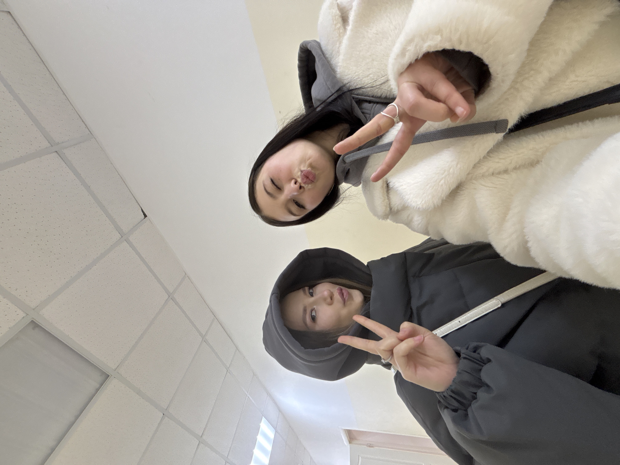 Two young women are posing for a selfie in an interior space with a drop ceiling. The woman in the top of the frame wears a white, furry jacket with a grey hood underneath, and is making a kissing face with her eyes closed, while holding up two fingers in a peace sign. The other woman, in the lower part of the frame, wears a black hooded puffer jacket and is also holding up two fingers in a peace sign with a slightly amused expression. The location appears to be indoors, likely a school, office, or public building given the presence of a drop ceiling with fluorescent lighting. No location data is available to infer the exact location in the city of Chaikovsky, Russia.