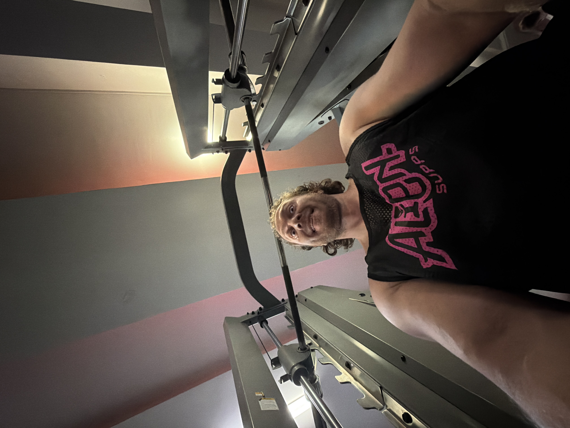 A man with curly brown hair and a wide, excited expression is captured from a low angle, looking up into a gym's Smith machine. He is wearing a black tank top with pink lettering that reads "ALON SUPPS". The man's arms are extended, and he appears to be either holding onto or interacting with the machine's bar. The background consists of the gym equipment and a ceiling with pink and gray panels, illuminated by a warm, artificial light source. The scene takes place inside a gym in Saint Petersburg, Russia.