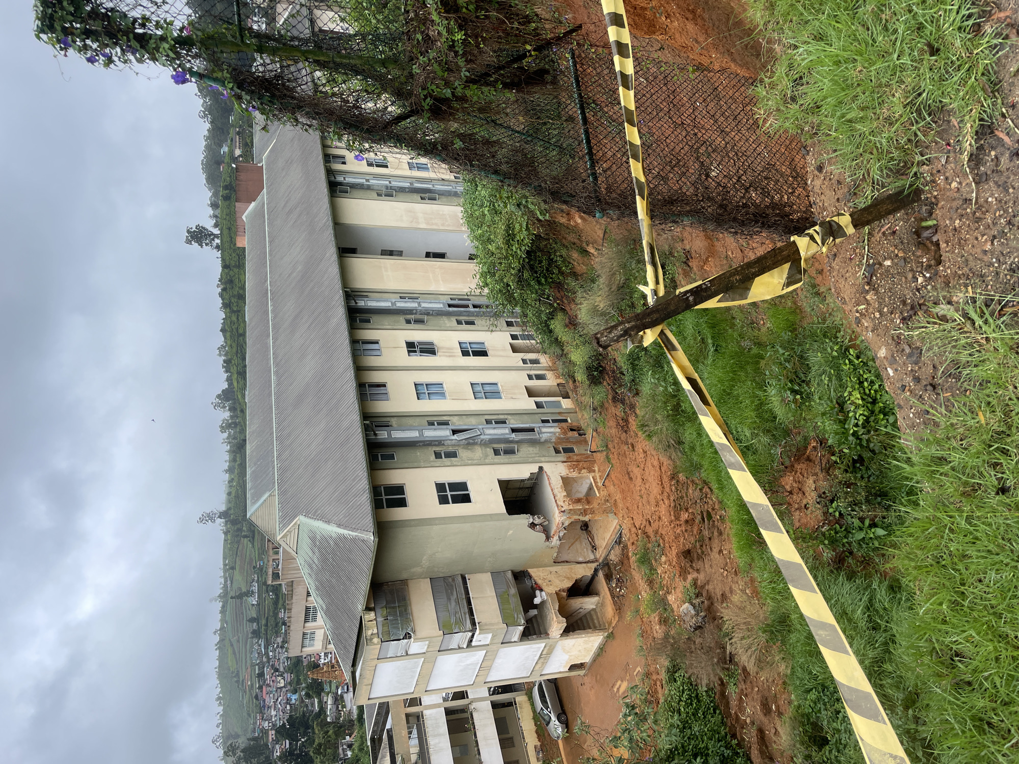 The image depicts a building in Nuwara Eliya, Sri Lanka, that has sustained significant damage, likely due to a landslide. The building's facade shows collapsed sections, exposing the interior structure. A silver car is parked in front of the building. Yellow and black caution tape is visible near the edge of the damaged area, suggesting a hazardous zone. The surrounding environment includes foliage, a chain-link fence, and a steep, bare earth slope, indicating the landslide's path. A town and terraced hills are visible in the distance. The sky is overcast, suggesting wet weather conditions.