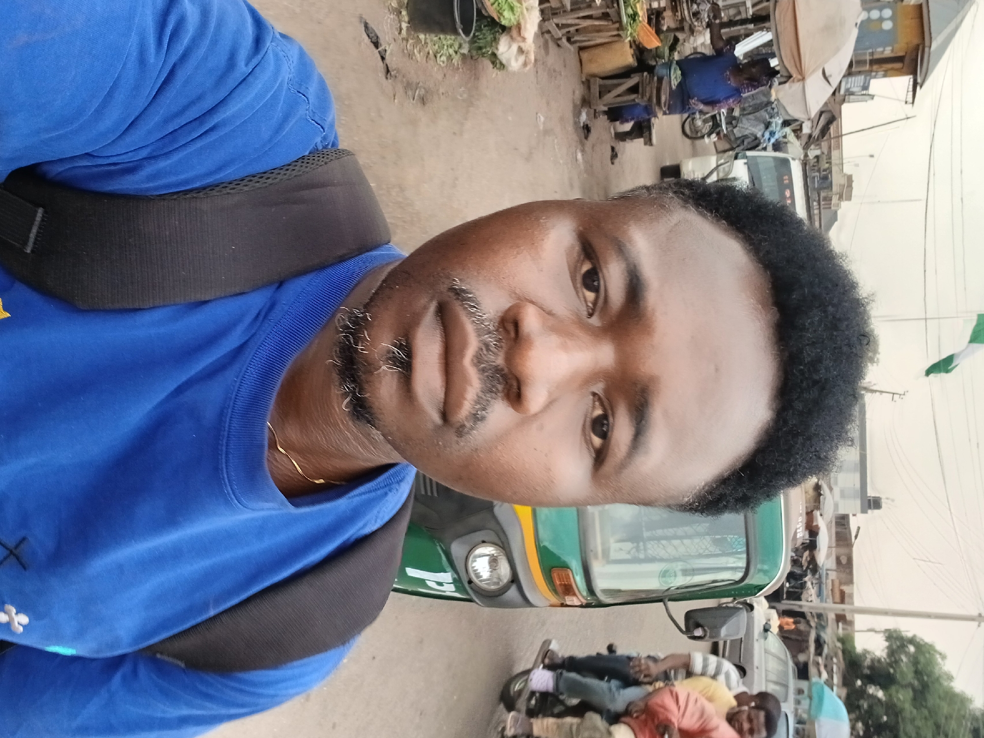 A medium shot of a man with dark skin, a black beard with some gray hairs, and short curly black hair wearing a blue t-shirt and a black backpack. He is looking directly at the camera.
Behind him is a green and yellow auto rickshaw, also known as a "keke" in Nigeria.
The background reveals a busy street scene, likely a marketplace. There are people on motorcycles, vendors with stalls, and various structures. A Nigerian flag is visible in the background. This scene is consistent with a street in Jos, Nigeria.