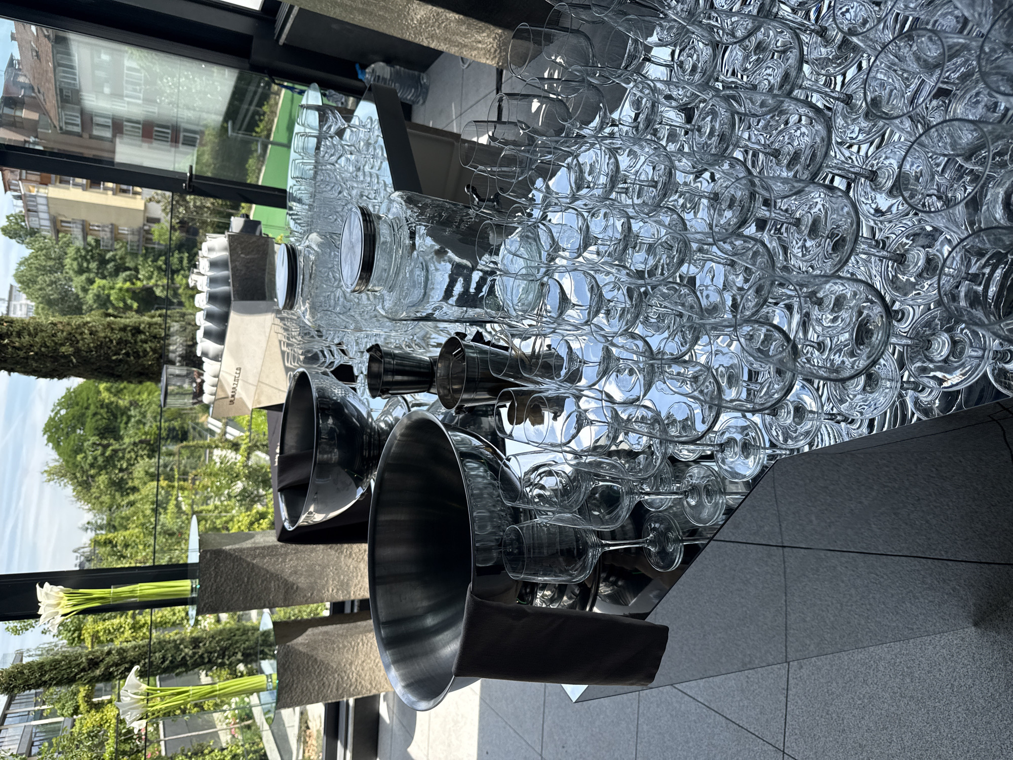 The image captures an indoor setting, likely a bar, restaurant, or event venue in Sofia, Bulgaria, looking out through a large window. The scene is dominated by a meticulously arranged display of glassware, primarily clear wine glasses and champagne flutes, inverted and stacked on a highly reflective surface, possibly a mirror or polished metal countertop. This arrangement creates numerous shimmering reflections, adding depth and a sense of luxury.
Among the glassware, several large, clear glass bottles or jars, one with a silver lid, are visible. To the left, large, polished silver or stainless steel bowls and smaller metal shakers are also part of the setup. One of the larger metal bowls has a dark fabric draped over its edge. In the foreground, white calla lilies with long green stems are arranged in slender vases, reflecting on a polished surface below them. The floor visible in the lower right is composed of gray tiles.
Through the large window, a bright daytime sky with some clouds is visible, along with the exteriors of several multi-story buildings, some with red-tiled roofs and balconies, typical of urban architecture. Lush green trees and foliage are abundant, suggesting a well-landscaped area outside. No people are present, indicating either an early setup for an event or a quiet period. The overall impression is one of cleanliness, readiness, and preparation. Partially visible text, possibly "MARIZZIA", can be seen on a box or structure in the middle ground, reflected in the surface.