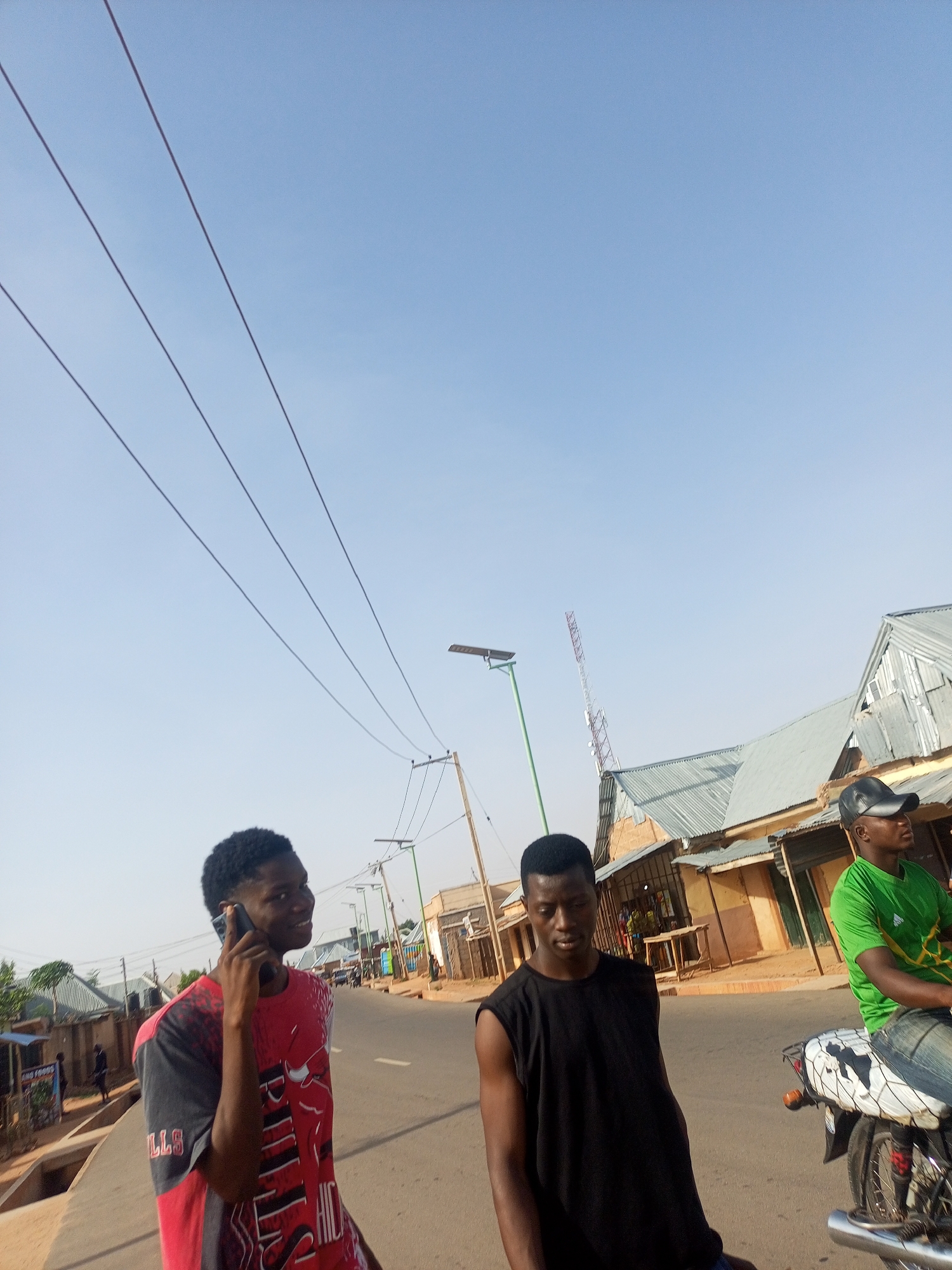 The image depicts a street in Tempure, Nigeria. Three young men are visible. One, wearing a red and black Bulls t-shirt, is on a mobile phone. Another, in a black sleeveless shirt, stands next to him. The third man, wearing a green t-shirt and a black cap, is seated on a motorcycle. The street is lined with buildings, mostly with corrugated metal roofs. There are utility poles and power lines overhead. A cell tower is visible in the background. The sky is clear and blue.