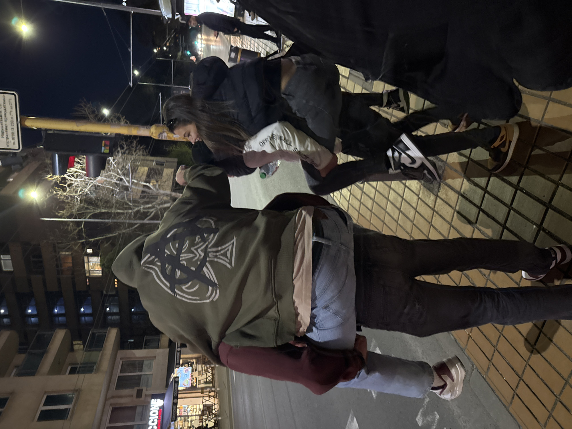 The image shows a group of people standing on a sidewalk at night. The location is identifiable as Sofia, Bulgaria, based on the presence of a sign written in Bulgarian and the distinctive architectural style of the surrounding buildings.
A woman in a dark jacket is hugging a person wearing a green hoodie with a large white graphic on the back. Other individuals are present in the frame, partially obscured. The surrounding environment includes streetlights, buildings, and signage. The sidewalk is paved with patterned tiles.