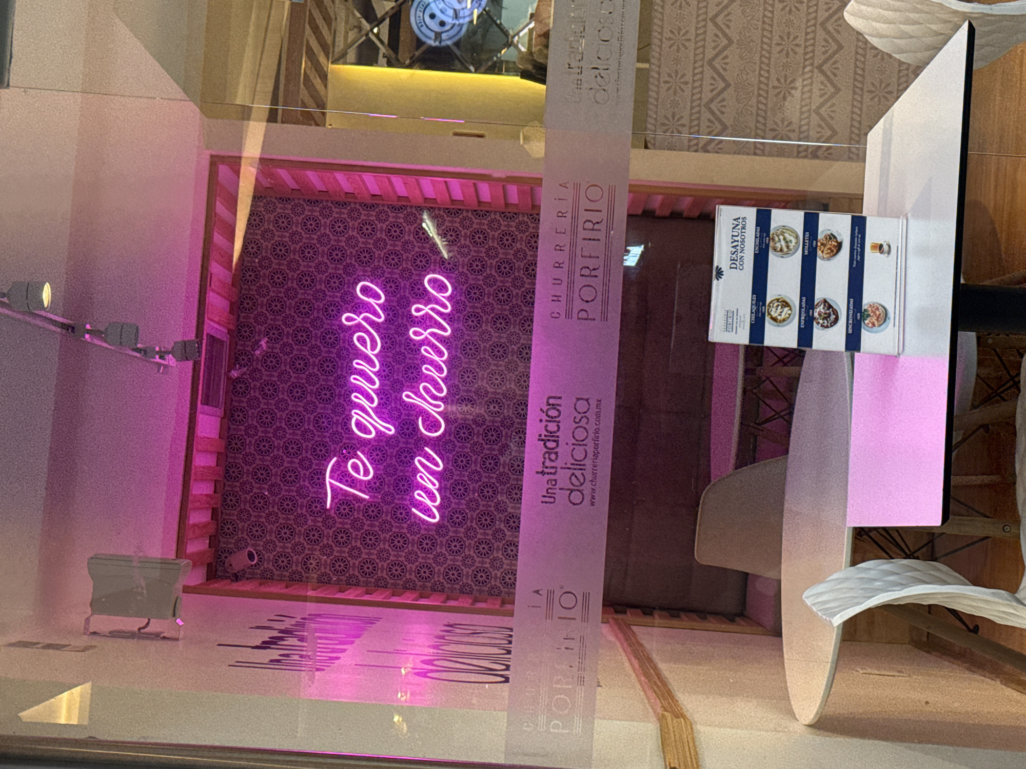 This indoor shot depicts the interior of a restaurant named Churrería Porfirio, located in the city of Puebla, Mexico. The scene is captured through a reflective glass or window. The restaurant's name is prominently displayed on a vertical banner alongside the phrase "Una tradición deliciosa" and its website. Inside, a neon sign in cursive Spanish spells out "Te quiero un churro", which translates to "I want a churro".
The restaurant's interior features a tiled wall with a repeating circular pattern. A menu stand displays breakfast options under the heading "DESAYUNA CON NOSOTROS" (Breakfast with us), listing dishes such as Chilaquiles, Enchiladas, Molletes, and Sincronizadas. In the foreground, a table with white chairs is visible, indicating a seating area. The environment suggests a modern yet traditional Mexican eatery specializing in churros and breakfast items.