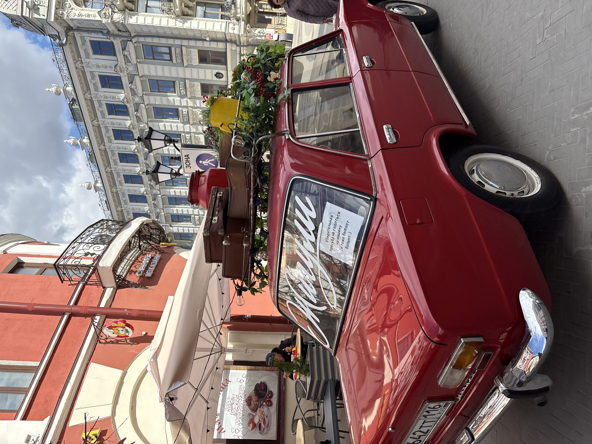 The image captures a red vintage car, likely parked as a display or attraction, in Kazan, Russia. The car has luggage and a yellow flower pot strapped to its roof. A sign in the car window, written in Russian, requests people not to sit on the car. The license plate is partially visible, and reads "642TT116 RUS".
In the background is a building with detailed facade ornamentation. To the left, a cafe or restaurant with an awning is visible. There's a menu board displayed with pictures of food. A person is seated at one of the tables. There is a sign that says "Из Казани" which translates from Russian to "From Kazan". A street sign that says "30HA" is visible to the left of the luggage. The overall setting suggests a tourist area or a street with restaurants and shops.