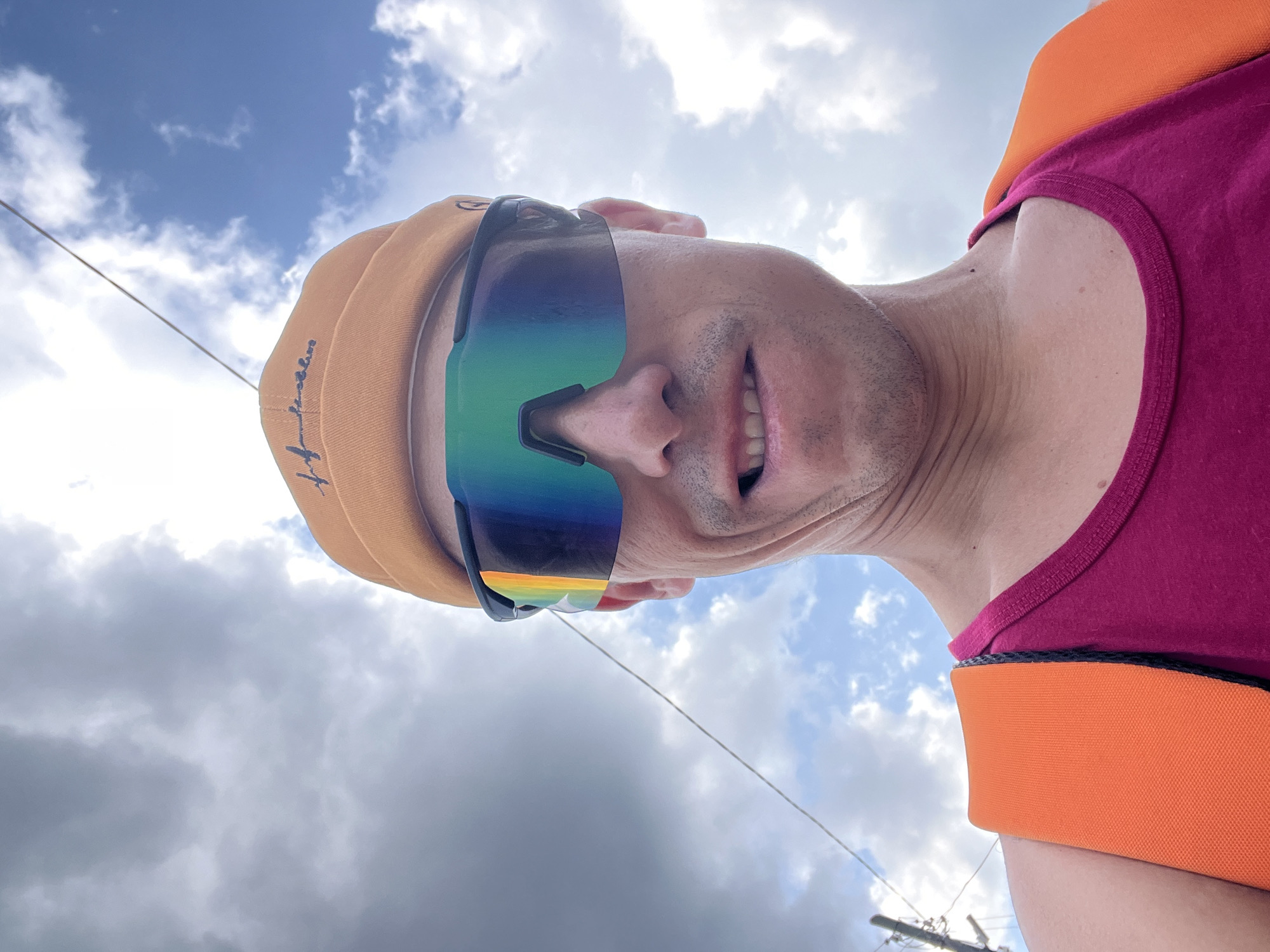 A person is taking a selfie outdoors. They are wearing an orange beanie and mirrored sunglasses. Their expression is one of mild grimace. They are wearing a pink tank top with orange straps over their shoulders. The background is a cloudy sky. The image does not contain any information that could be used to determine the location of Chaikovsky, Russia.