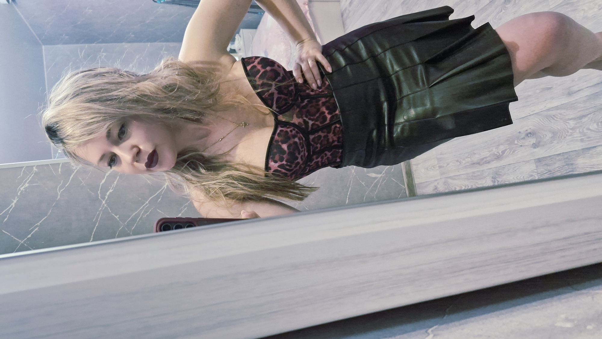 A woman is posing for a photo in a bathroom. She's wearing a leopard print top and a black leather skirt. The bathroom has gray marble walls and a light wood-patterned floor. The woman is holding a red phone in her left hand, presumably taking the photo in a mirror.
I cannot verify the location in the city of Otchor, Russia.