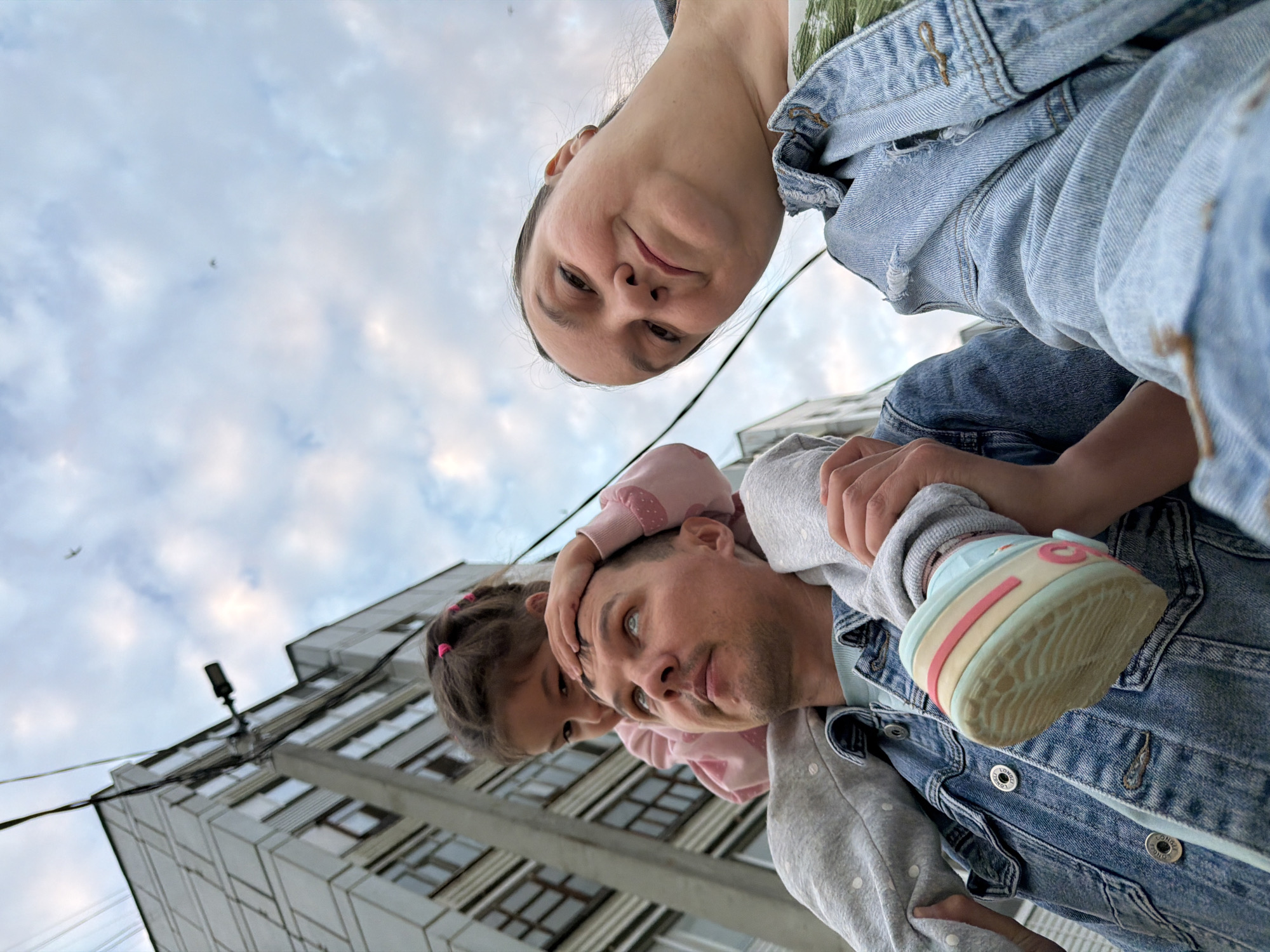 A low-angle, wide shot captures a family of three outside a building in Chaikovsky, Russia. In the foreground, a father is carrying a young daughter on his shoulders. The father is wearing a denim jacket and a light blue t-shirt. The daughter is wearing a pink sweatshirt and a grey hoodie, and has her hand on her father's head. To the right of the father, a woman, presumably the mother, is looking at the camera and smiling. She is wearing a denim jacket and a green top. Behind the family, a multi-story apartment building is visible against a cloudy sky. A bird is seen flying in the distance. The image appears to be a selfie taken by the mother or father.