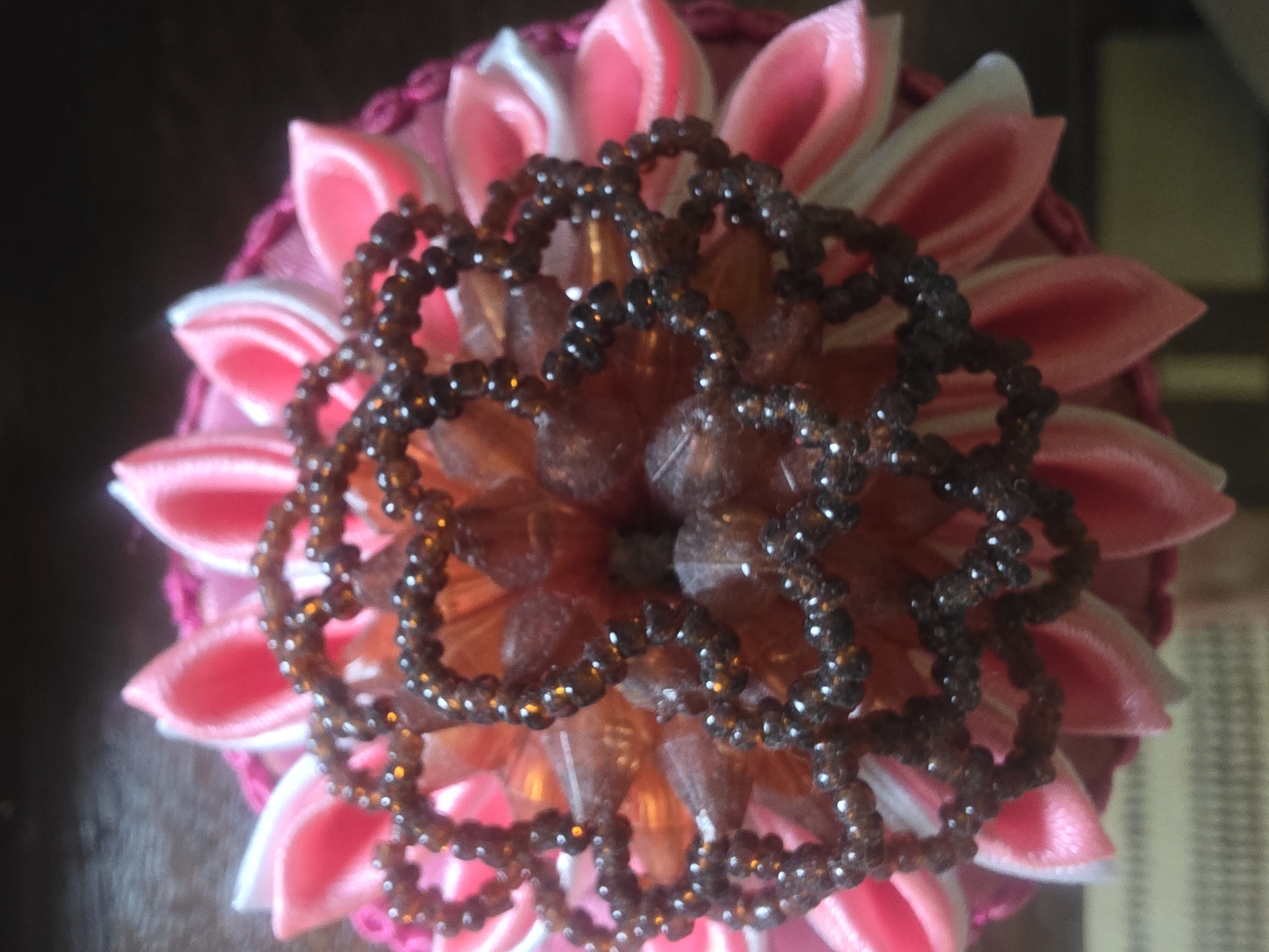 The image shows a close-up, overhead view of a decorative flower craft, likely a hair accessory or embellishment. The flower features multiple layers of satin petals, primarily in shades of bright pink and white, arranged in a radial pattern. At the center of the flower is an intricate, layered embellishment made of small, iridescent brown beads, forming a delicate, web-like structure. The background appears to be a dark, textured surface, possibly wood. There are no people or animals visible in the image. The specific location within Bila Tserkva, Ukraine, is not discernible from the visual information provided.