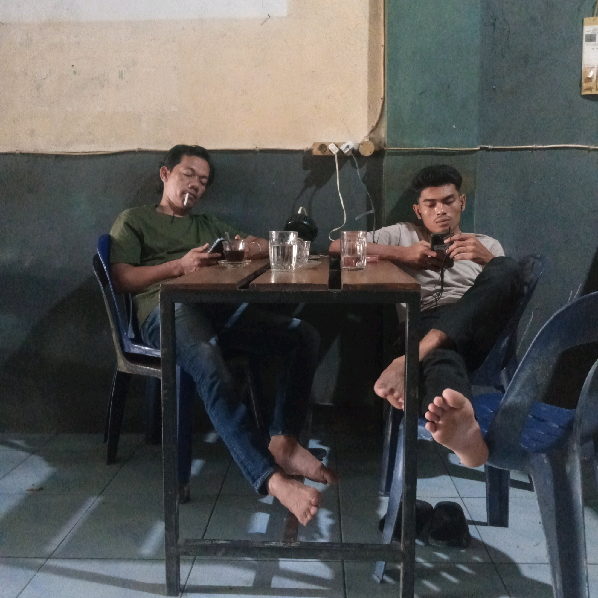 Two men are seated at a wooden table in what appears to be an outdoor or semi-outdoor establishment, possibly a warung kopi, in Grong-Grong, Indonesia. The man on the left, wearing a green t-shirt and blue jeans, is smoking a cigarette and looking at his smartphone. He has a glass of dark liquid, likely coffee, on a saucer in front of him. The man on the right, wearing a light grey t-shirt and black pants, is also looking at his smartphone, and has earbuds in. There is a glass of clear liquid in front of him, likely water. The table is set on a tiled floor, and there are two blue plastic chairs visible. The lighting suggests it is either evening or a dimly lit interior. The background consists of a textured wall and some electrical outlets. The overall atmosphere is relaxed and casual.