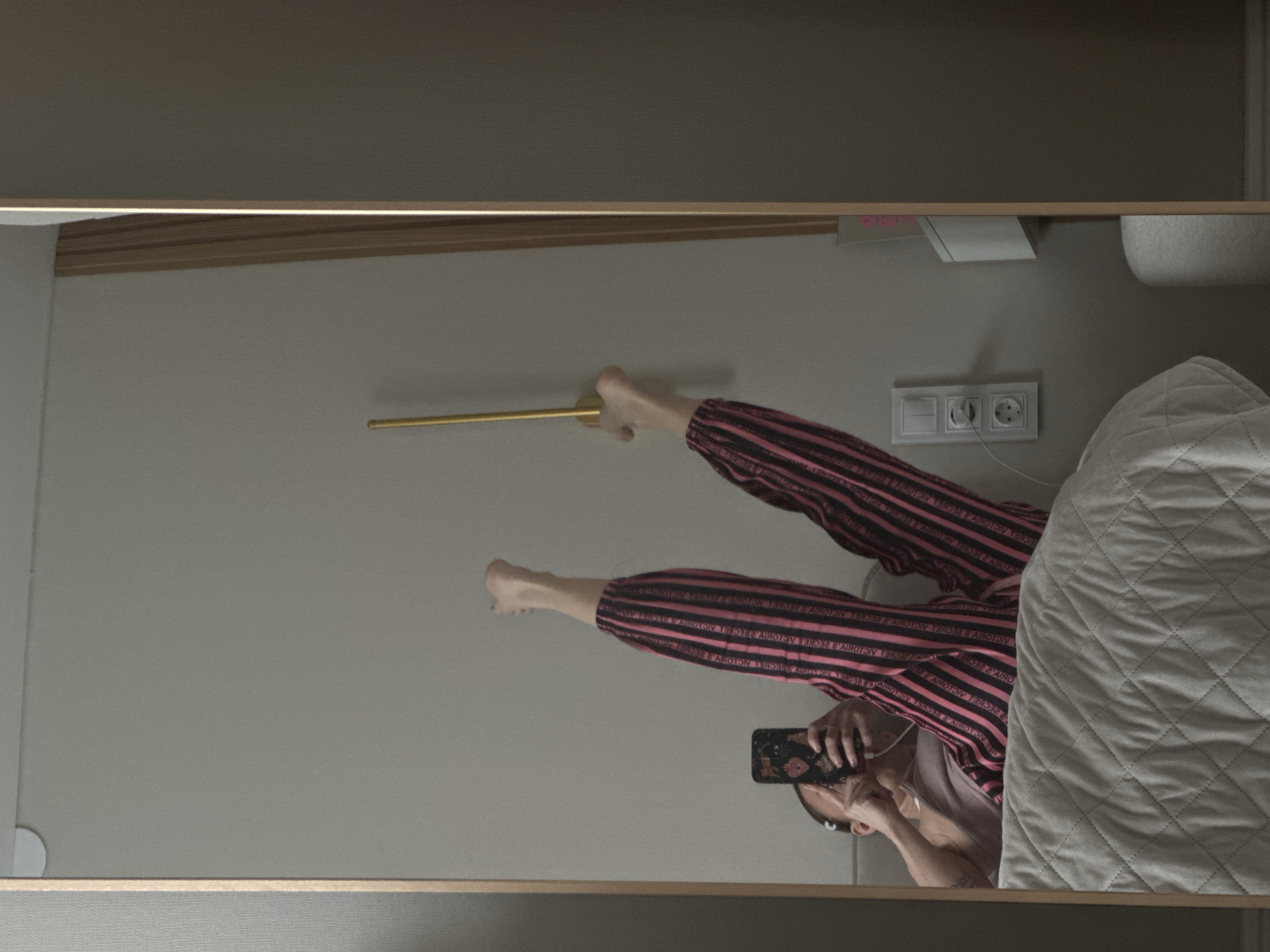 A reflection in a mirror shows a person in pink and black striped pajamas lying on their back on a bed. The person is holding a phone and appears to be taking a photo in the mirror. Next to the bed, there's a white comforter. A golden metal rod extends along the wall above the bed, and an electrical outlet is visible on the wall above the bed. This image does not contain enough information to establish a location in the city of Votkinsk, Russia.