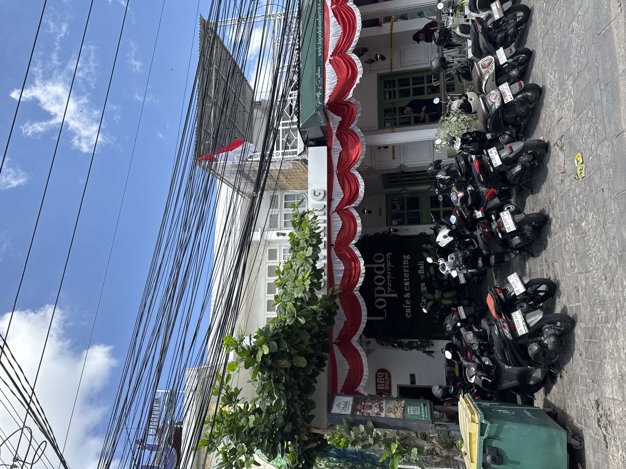 A street-level outdoor view of a commercial building in North Kuta, Indonesia, features a facade decorated with red and white fabric drapes. A sign above the entrance reads " Lopodo cafe & catering". Parked motorcycles are lined up in front of the establishment, and a large green dumpster is situated to the left of the frame. Overhead, a complex network of electrical wires crisscrosses the sky, which is partly cloudy. A red and white Indonesian flag is visible on the roof of the building.