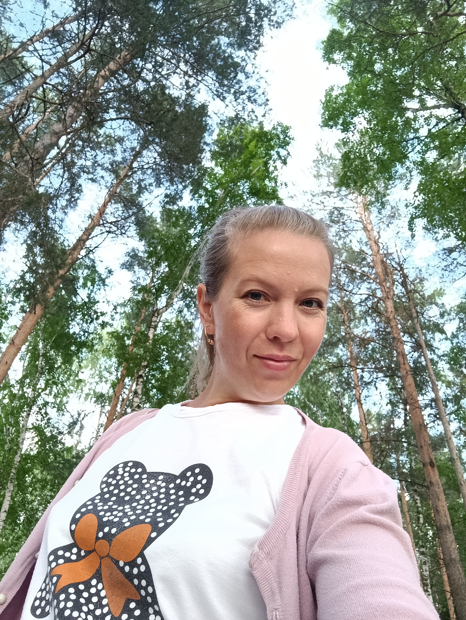 A low-angle, medium shot captures a woman taking a selfie in a forest with tall trees and a bright sky. The woman, positioned in the lower right of the frame, faces the camera with a slight smile. She has blonde hair pulled back and is wearing a white t-shirt with a black and white polka-dot teddy bear graphic and an orange bow, layered under a pale pink cardigan. The background consists of a dense forest with slender trees, some with light bark and others with darker trunks, interspersed with green foliage. The upper portion of the image is dominated by the sky, which appears partially overcast with bright patches. The scene suggests an outdoor setting, possibly a park or natural area, likely within the city of Chaikovsky, Russia, given the context provided.