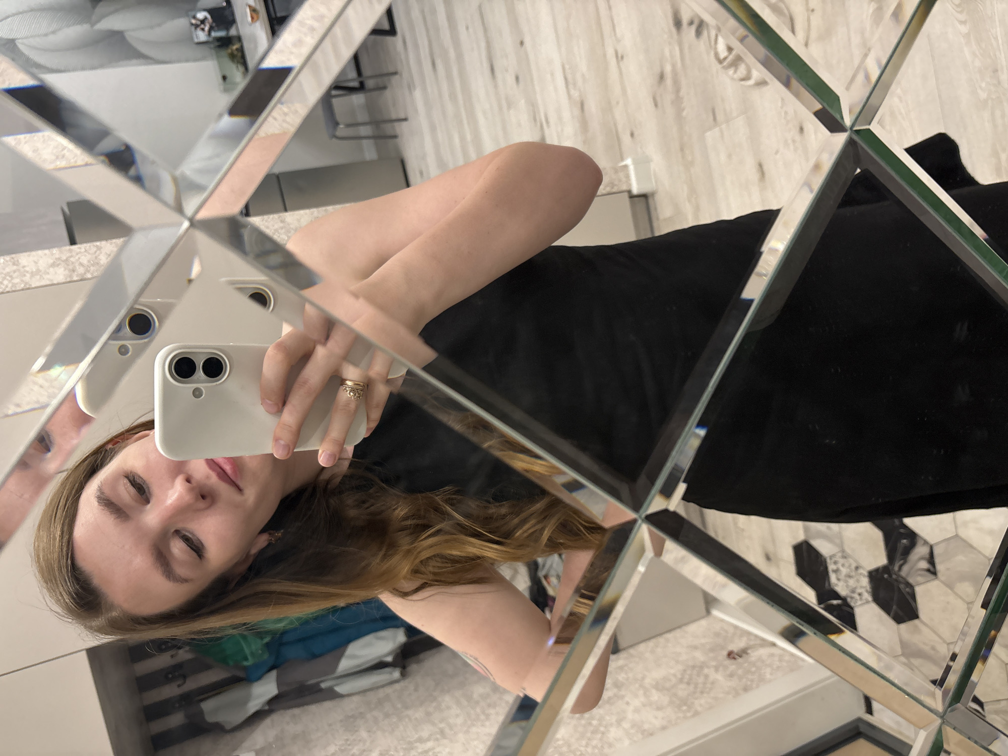 The image depicts a woman taking a selfie in a mirror composed of multiple triangular segments. She is holding a white smartphone. The woman has light skin, brown hair, and is wearing rings on her fingers. She appears to be wearing a black dress. The environment reflected in the mirror includes flooring with a geometric pattern, possibly in a bathroom or dressing area. Based on available information, the location in the city of Izhevsk, Russia cannot be determined from the visual content of the image.