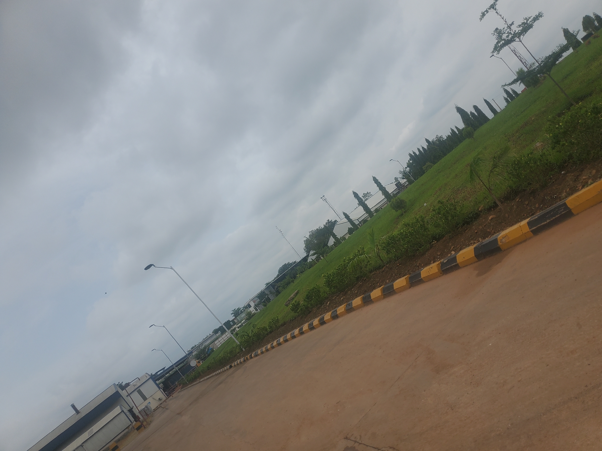 The image captures a tilted view of a paved road in Gwagwada, Nigeria, bordering a meticulously maintained green lawn. The lawn is densely covered with grass and punctuated by scattered shrubs and young trees. A yellow and black striped curb demarcates the edge of the road from the lawn. In the background, several modern buildings with white exteriors and dark roofs are visible, suggesting an industrial or commercial area. Tall, slender streetlights stand along the road. The sky is overcast with heavy gray clouds, indicating a cloudy day. The overall impression is one of an orderly and developed urban or peri-urban environment.