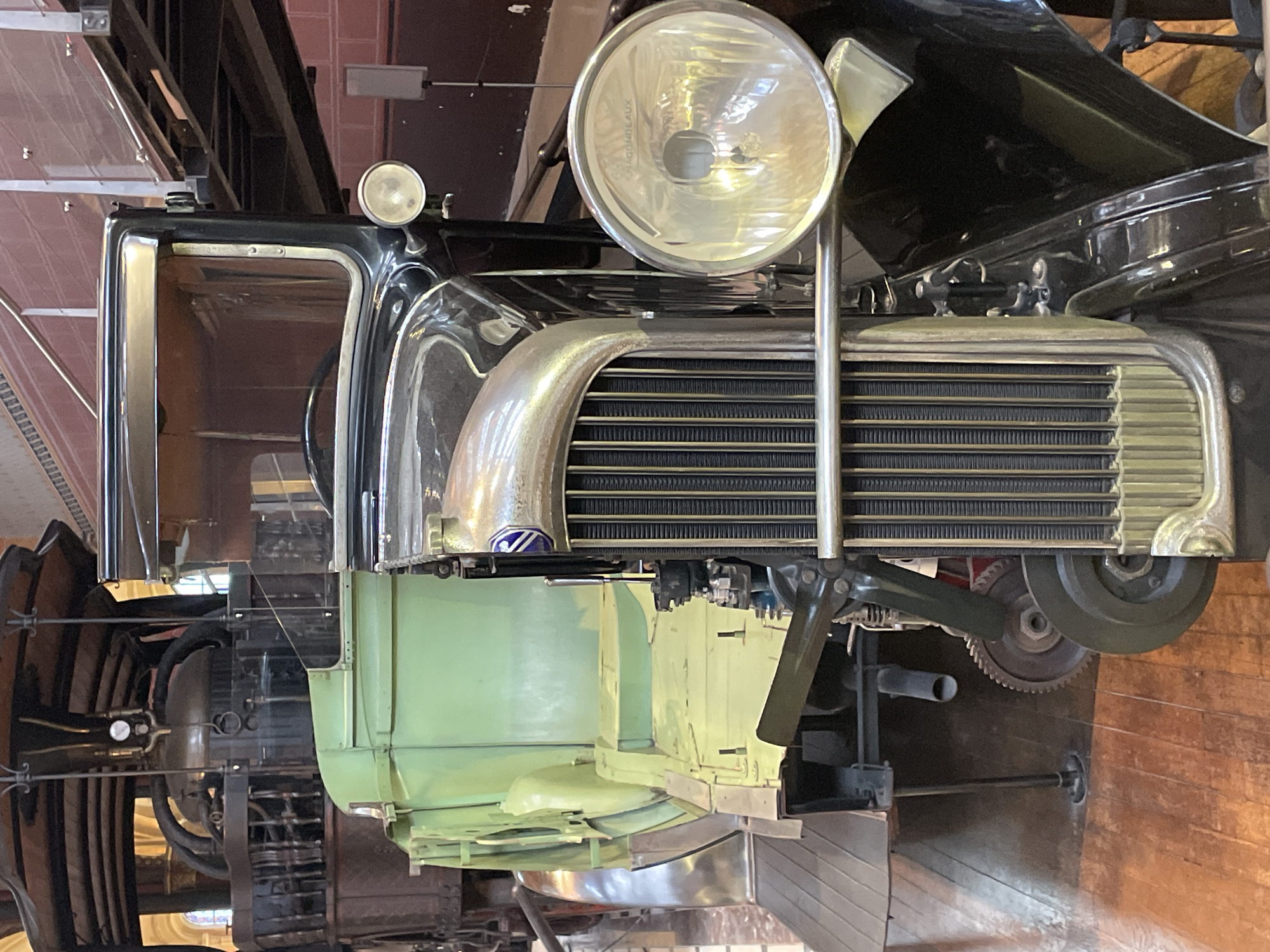 The image shows a close-up, head-on view of the front section of a vintage truck, possibly on display. The truck is predominantly black with a bright green section visible behind it, likely the cargo bed or body. The front features a prominent, ribbed chrome grille and a large, circular headlight on the right side. The truck appears to be indoors, as indicated by the polished wooden floor and the indistinct background which includes architectural elements like a railed walkway and what seems to be part of another vehicle or machinery. The lighting is bright, highlighting the shiny surfaces of the truck. The context of Paris, France, is not evident from the visual information in the image itself.