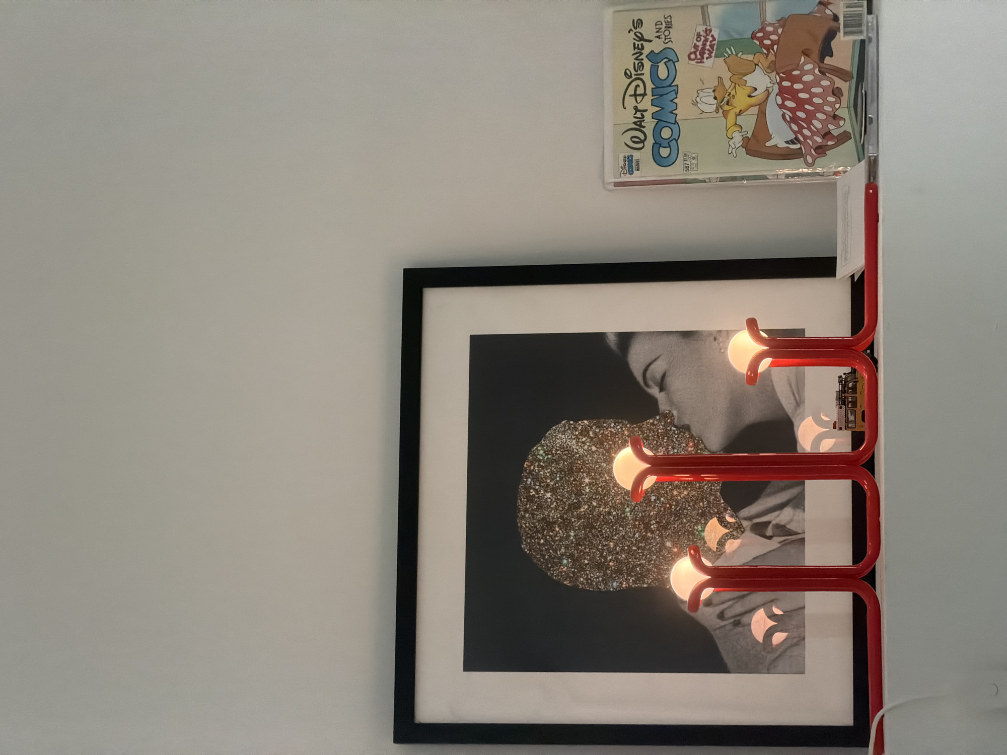 The scene presents a close-up, interior view of a wall adorned with various objects. A framed black and white photograph depicting a person kissing a glittering shape takes prominence. A red lamp with multiple light bulbs is affixed to the wall alongside the picture. Above these, a Walt Disney's Comics magazine is placed on a shelf. The wall is painted in a light color. While the scene appears indoors, it doesn't offer enough details to pinpoint a specific location within Los Angeles, United States of America.