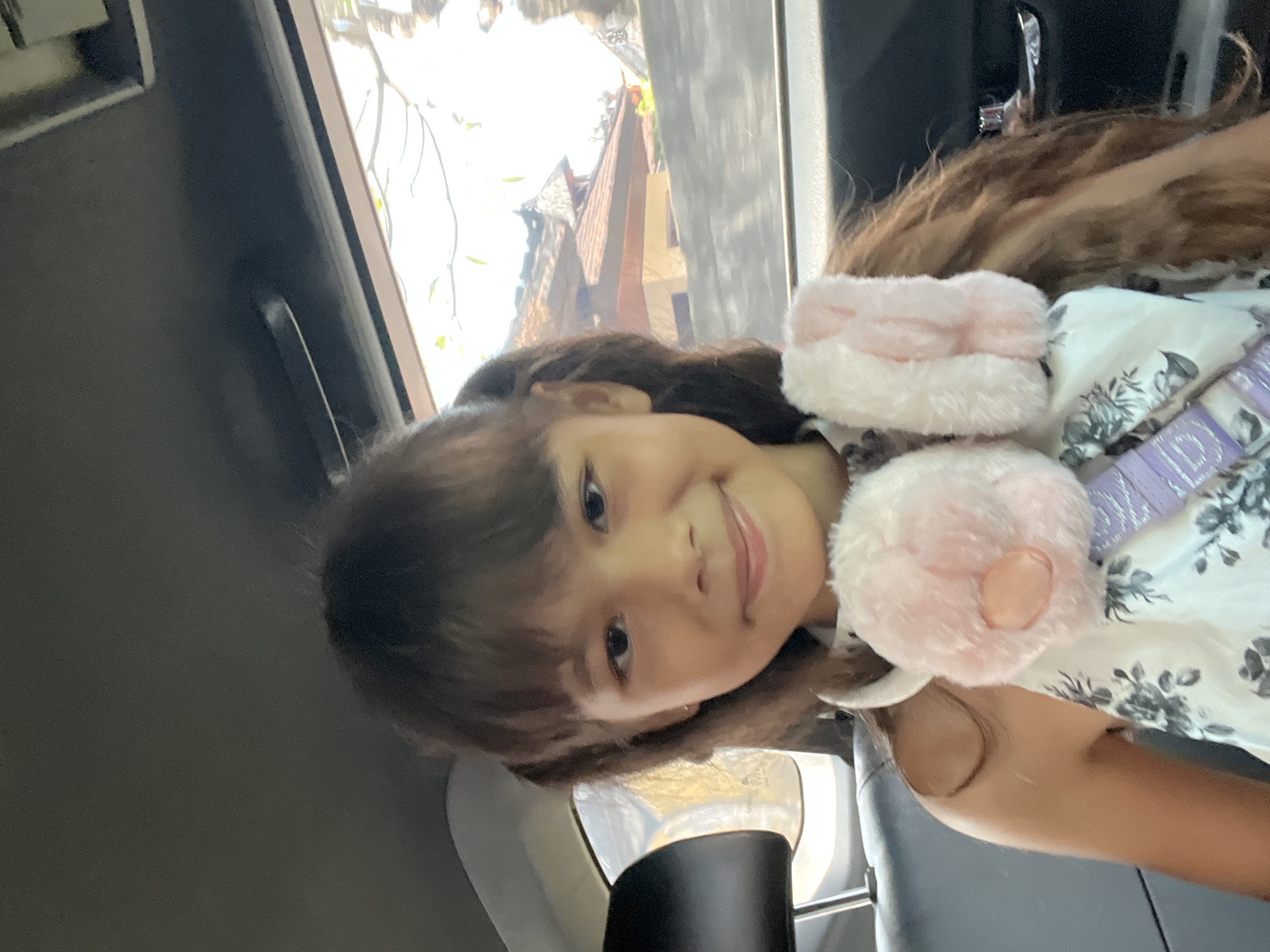A young girl with long, dark brown hair styled in pigtails is seen from the chest up, seated in the back of a vehicle. She is wearing a white dress with a floral pattern and has pink furry headphones around her neck, decorated with bunny ears. She is looking directly at the camera with a slight smile. Outside the car window, traditional Balinese architecture with tiled roofs is visible, suggesting the location is North Kuta, Indonesia. The lighting indicates it is daytime, with sunlight filtering through the window.