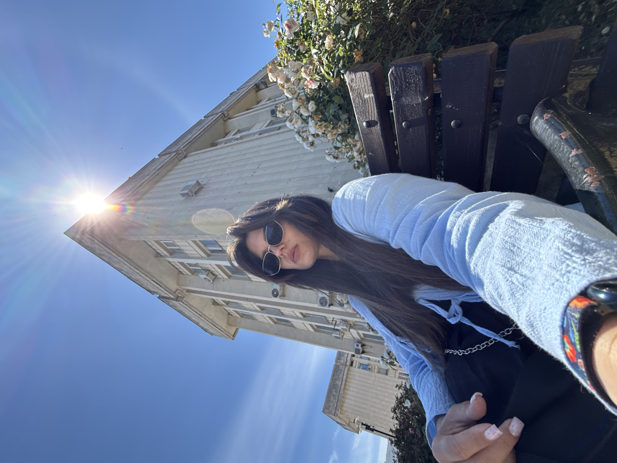 A woman with long dark hair and sunglasses sits on a wooden bench outdoors. She is wearing a light blue open-collared shirt over a black top, with a silver chain visible. Her left hand is resting on her lap, with manicured nails. Behind her, a modern building with multiple windows and air conditioning units rises into a clear blue sky. Sunlight is shining brightly from the top of the building, causing lens flare. To the right of the building, a bush with white and pink flowers is in bloom. The bench is dark brown and shows signs of wear. The overall scene suggests a sunny day in an urban or semi-urban setting.