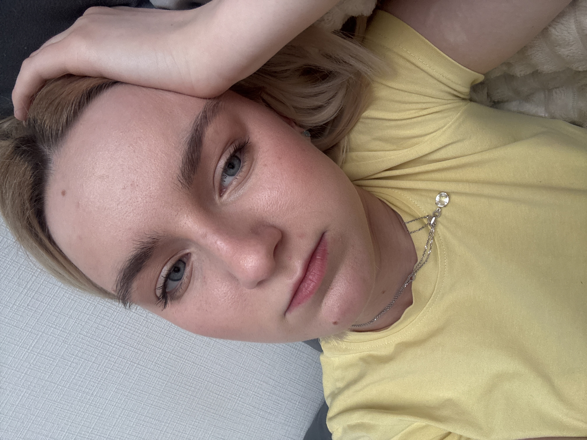 A young woman with fair skin, light blue eyes, and blonde hair lies on a gray textured surface wearing a yellow t-shirt with a silver necklace. Her left hand rests on her forehead. The photograph does not show enough information to determine if the image was taken in Vsevolozhsk, Russia.