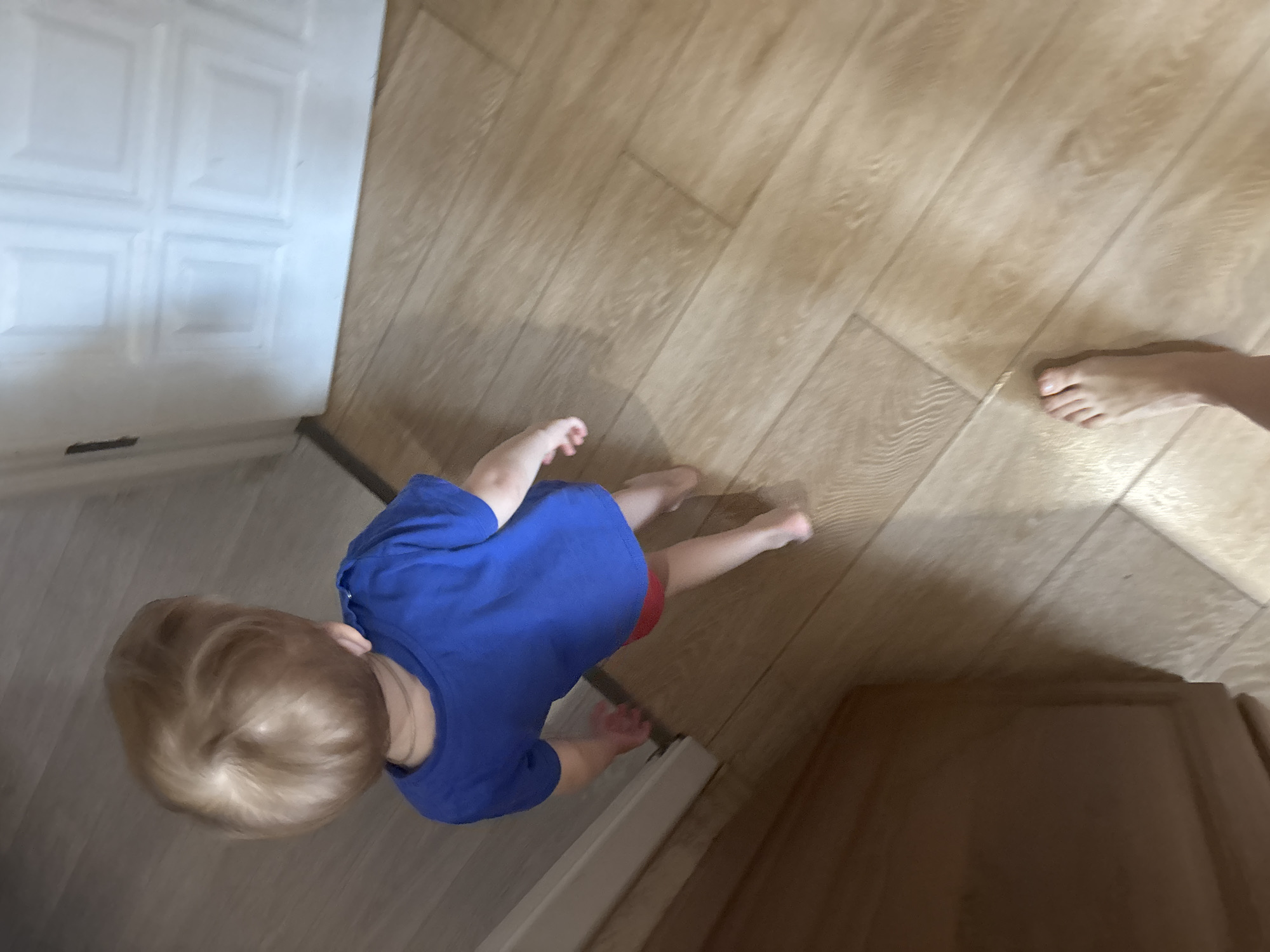 An overhead view captures a young child walking forward across a light-colored, wood-effect floor. The child, with light blonde hair featuring a prominent crown swirl, is wearing a royal blue short-sleeved t-shirt and red shorts, and is barefoot. A thin, light-colored chain is visible on the child's neck. To the right, a white paneled door with four recessed panels is visible. To the left, a portion of a dark brown or wooden furniture item or wall panel is present. In the lower right foreground, a bare adult human foot is partially visible, indicating the presence of another individual behind the child. The scene is indoors.