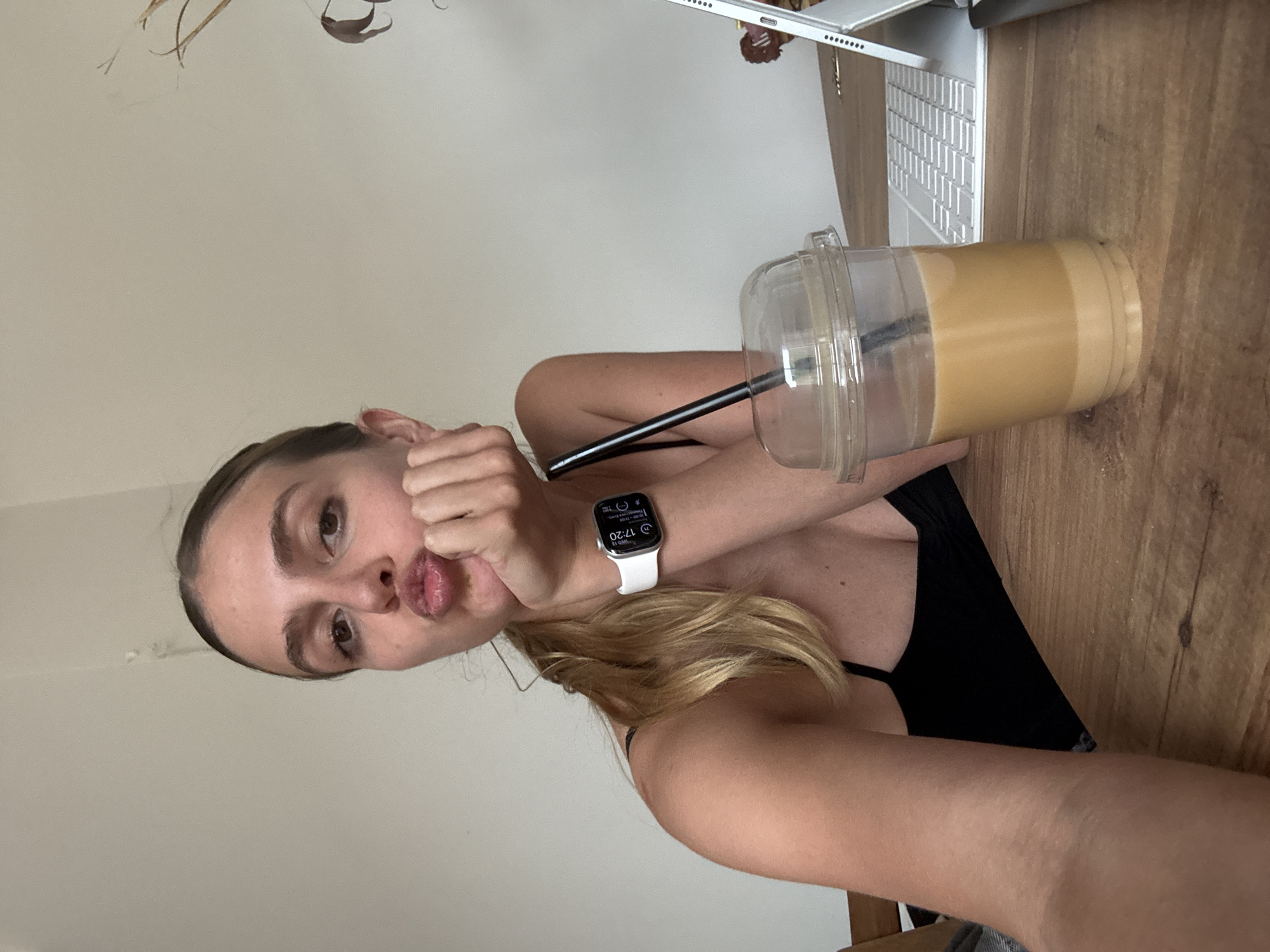 A young woman is sitting at a wooden desk with a laptop and a cup of coffee. The woman is wearing a black tank top and an Apple Watch on her left wrist. The laptop is open and the keyboard is visible. The cup of coffee has a straw in it. The desk is made of wood. The location in the city of Unknown, Unknown cannot be determined from the image.