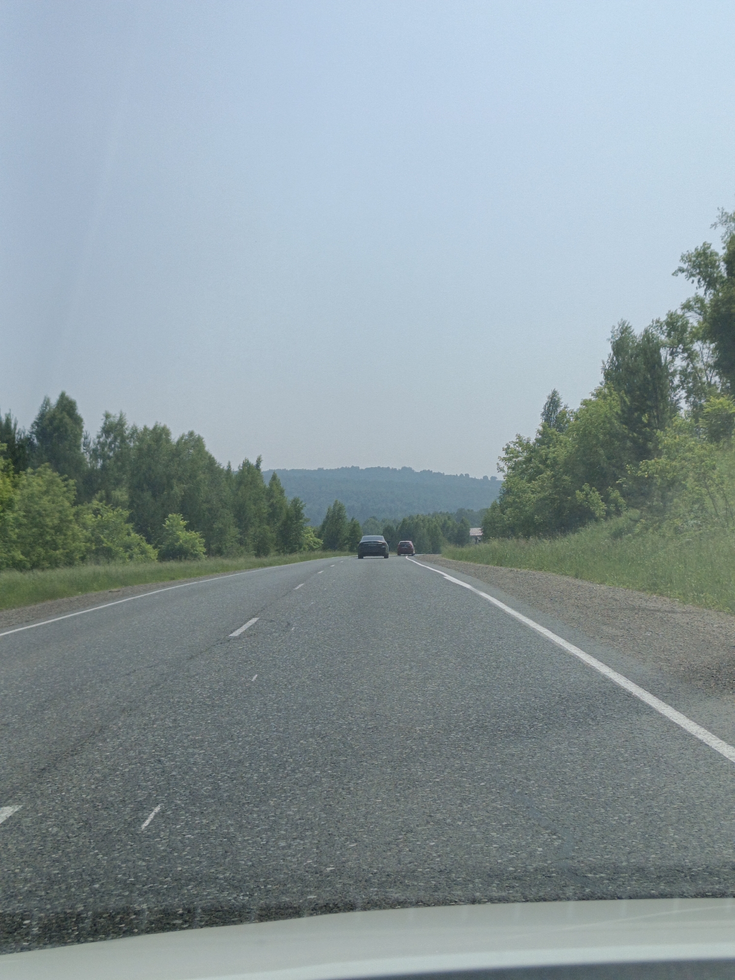 The image depicts a view from a vehicle driving on a two-lane asphalt road. The road has white dashed lines indicating lanes. On the left side of the road, there is a dense line of green trees and foliage. On the right side, the vegetation continues, and further in the distance, rolling hills covered in trees are visible. The sky is overcast with a pale blue hue. Two cars are visible in the distance on the road, one dark-colored and one red. The perspective suggests the photo was taken from the driver's or front passenger's seat of a moving vehicle, with the dashboard visible at the bottom of the frame. The overall environment is rural and appears to be a highway or a major road passing through a wooded, hilly landscape.