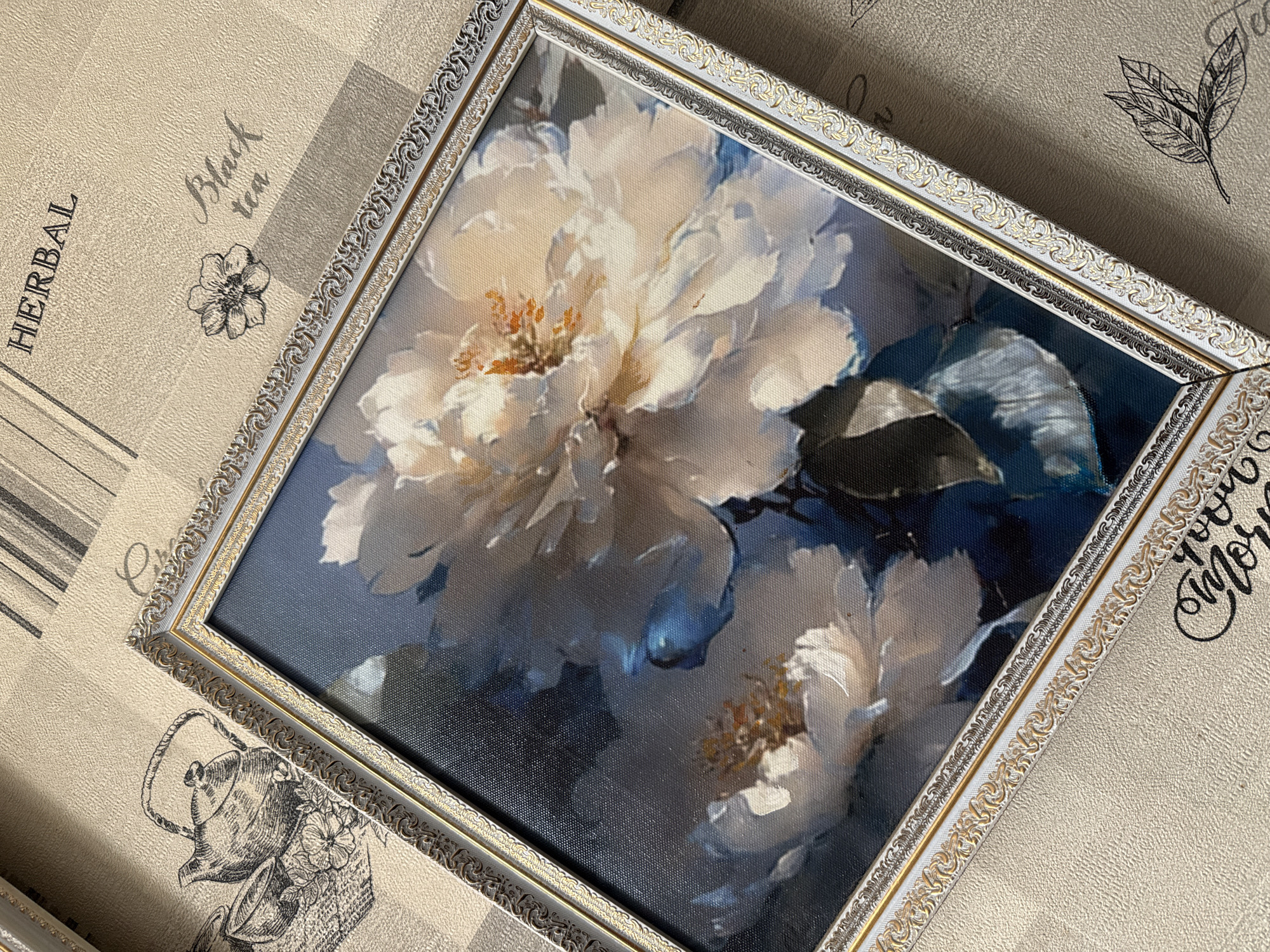 The image shows a framed painting of white peonies on a textured, light-colored surface. The painting is positioned at an angle, and its ornate frame is visible, featuring white and gold detailing. The background surface has decorative elements, including a sketch of a teapot and teacup on the left, and text that appears to read "HERBAL" and "Black tea" in the upper right portion. There is also cursive text at the bottom that is partially obscured. The lighting suggests an indoor setting, possibly on a table. There is no indication of the location being Simferopol, Ukraine.
