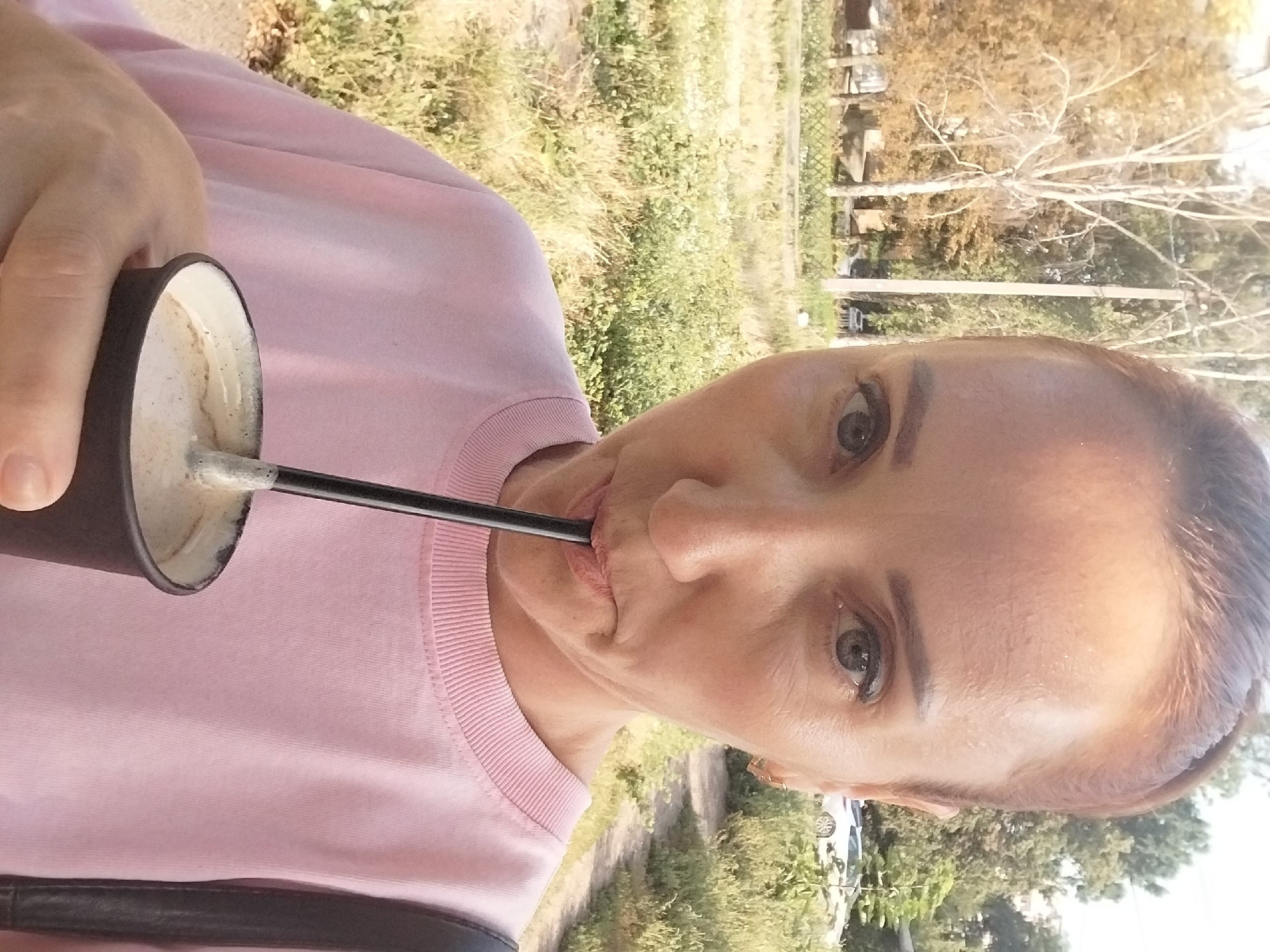 A woman with auburn hair pulled back in a ponytail, wearing a light pink sweatshirt, takes a sip of a beverage from a dark brown paper cup with a black straw. She is outdoors, with a background of trees and dry grass, and a dirt path. A white car is visible in the distance. The setting appears to be an urban or suburban area.