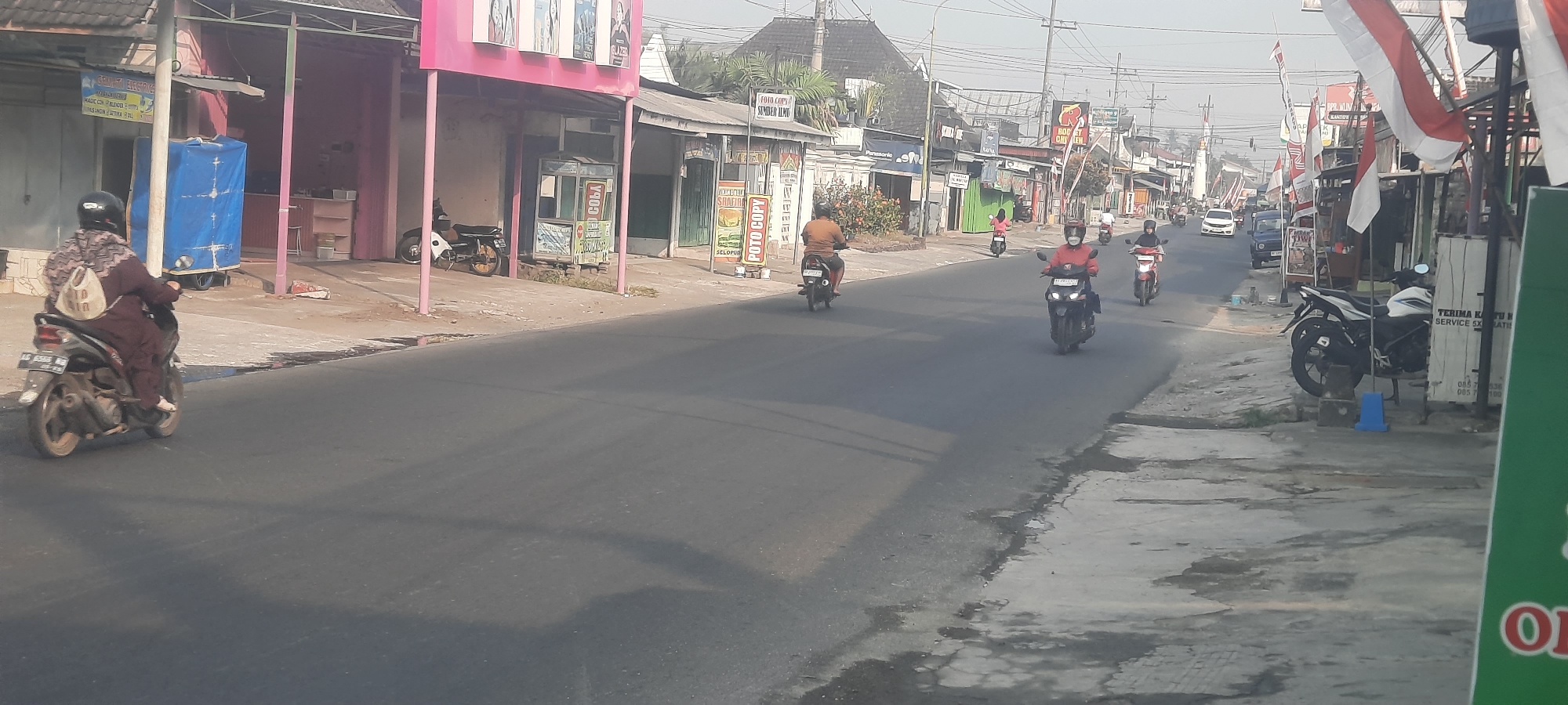 This is a street-level, medium-shot photograph taken on a street in Selopuro, Indonesia. The scene depicts a roadside environment with several motorcycles and a few cars. The sky is overcast, and the overall lighting suggests it is daytime.

On the left side of the street, there are several buildings with storefronts. One building has a pink facade and a white awning. A person wearing a dark brown outfit and a helmet is riding a motorcycle on the left side of the road, facing away from the camera. The motorcycle has a license plate that reads "AG 6388 KG". Next to this building, a blue tarp covers an object on a cart. Further down the sidewalk, a white and black motorcycle is parked.

On the right side of the street, more buildings and storefronts are visible. Red and white Indonesian flags are displayed along the roadside. Several people are riding motorcycles on the street. One person in a red jacket is riding a dark-colored motorcycle. Another person in a red helmet is on a red motorcycle. Further down the road, a white car and a dark blue car are visible. A white and black motorcycle is parked on the right sidewalk.

The road surface appears to be asphalt, with some patches of water or stains visible on the right side. The environment suggests a commercial or residential area with moderate traffic.
