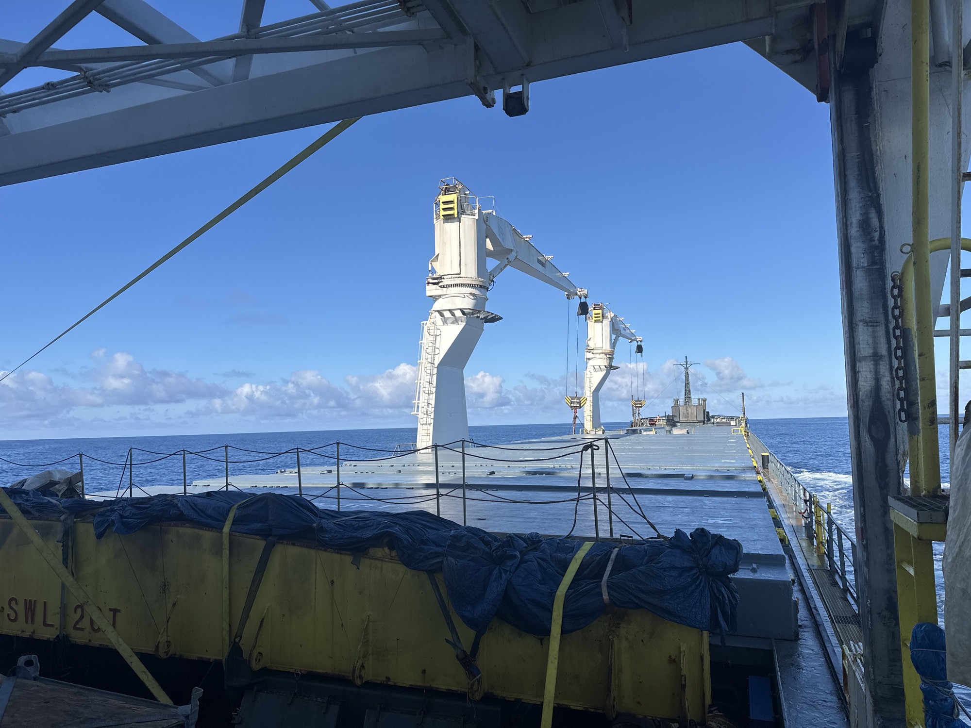 The image depicts the deck of a cargo ship at sea. Two large, white cranes are visible on the deck, along with a yellow container covered with a dark blue tarp secured with yellow straps. A railing runs along the edge of the deck. The sky is blue with scattered clouds, and the ocean stretches to the horizon. The location is unknown.