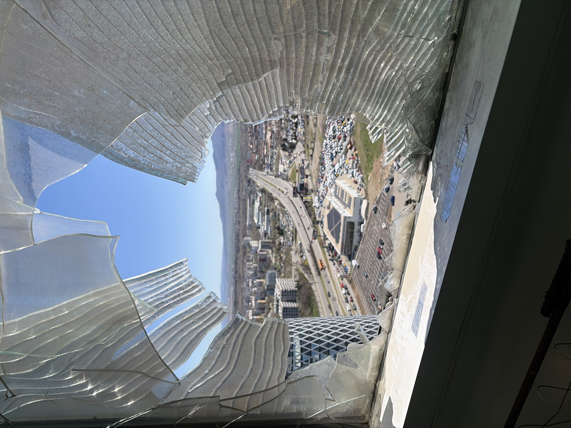 The image shows a view from a broken window of a high-rise building. The window glass is shattered with a hole in the middle revealing a cityscape. In the background, there are buildings, roads with moving cars, and a mountain range. In the foreground is the window frame and shards of glass, partially obscuring the view. A building with a diamond grid design is visible through the hole in the broken window. The location is most likely Sofia, Bulgaria.