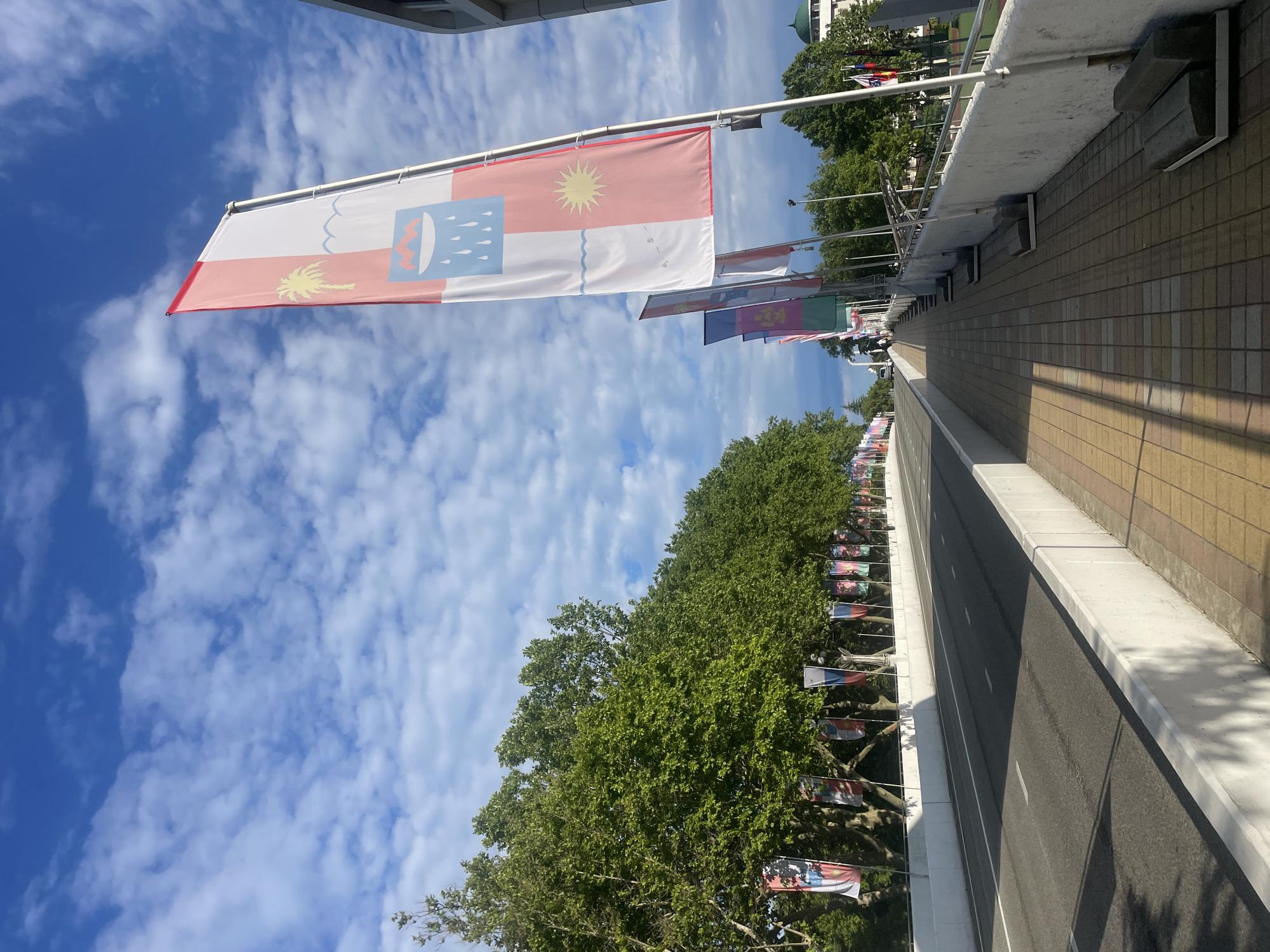 The image shows a street lined with flags on both sides, under a partly cloudy blue sky. The street appears to be a roadway for vehicles. On the right side, there is a pedestrian walkway paved with yellow and brown tiles, bordered by a white concrete barrier with metal railings. Benches are placed along the walkway. A row of trees with lush green foliage is visible on the left side of the road, also adorned with flags. The flags are vertical banners, with one prominent flag in the foreground featuring a red and white design with symbols of a palm tree, waves, mountains, and a sun. Further down the street, more flags are visible, suggesting a formal procession or event. In the distance, buildings with a green domed roof can be seen. This scene is likely located in Sochi, Russia, possibly near the Olympic Park, which is known for its grand displays of international flags.