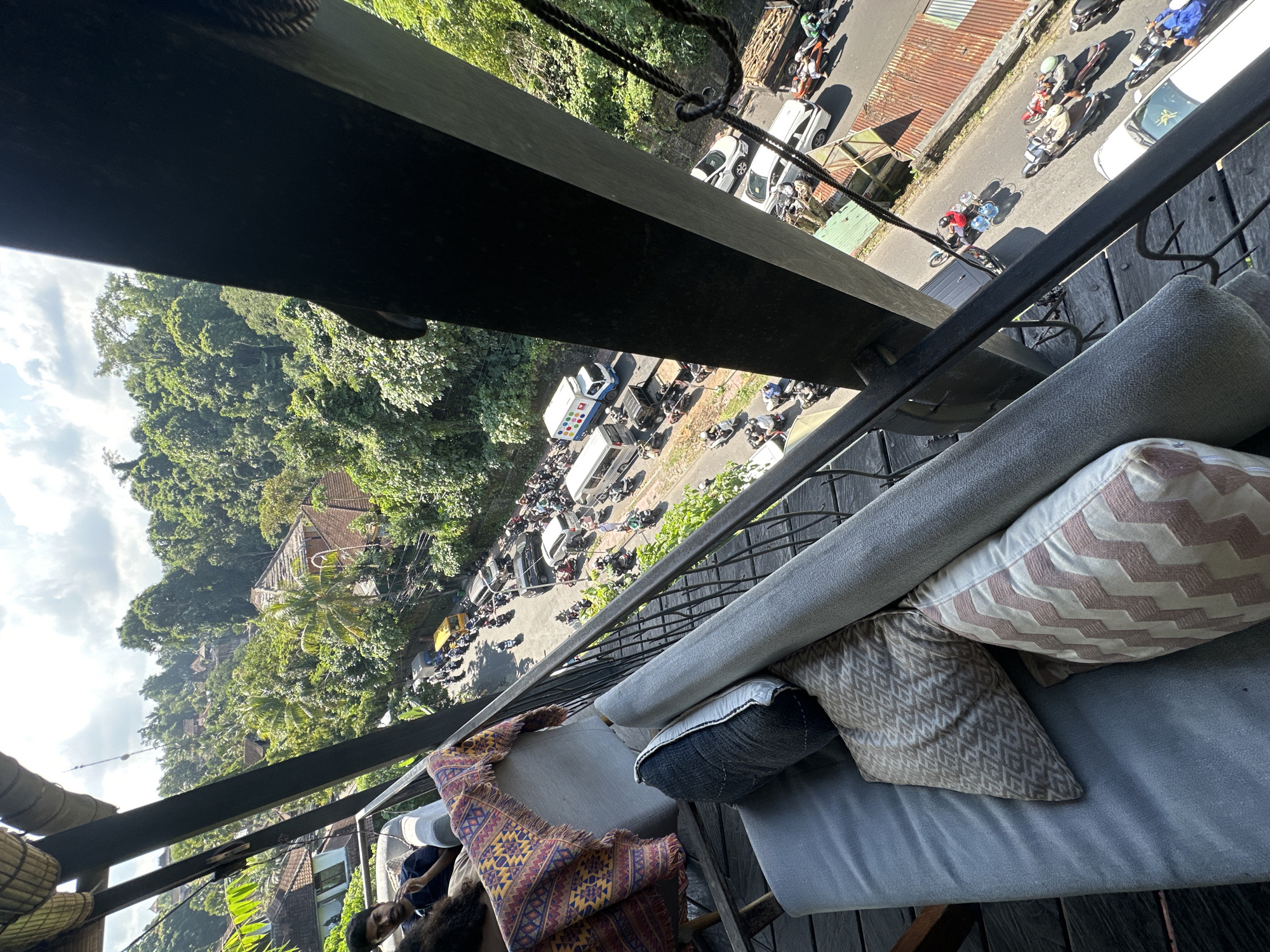 An elevated view from a balcony overlooks a traffic jam on a road lined with lush green trees and scattered buildings, suggesting a location in or near Ubud District, Indonesia. The road is congested with numerous motorcycles, scooters, cars, and a few vans, indicating heavy traffic. A yellow bus is visible among the vehicles. The environment appears tropical, with dense foliage and rooftops suggesting a residential or commercial area interspersed with natural greenery. Sunlight streams from the upper right, casting shadows and illuminating the scene. In the foreground, a portion of a balcony is visible, featuring a gray sofa with several pillows and a colorful patterned blanket draped over it. Two individuals are partially visible on the sofa, one looking away from the camera. The railing of the balcony creates a visual barrier, framing the view of the street below.