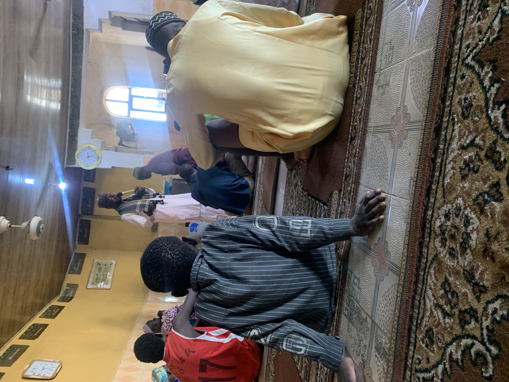 In a room with yellow walls and a wooden ceiling, several people are gathered, some seated on the floor on patterned rugs. A man in a light pink robe and a checkered scarf stands at the front, holding a microphone and speaking. To his left, a person wearing a red vest with the number "17" is seated. In the foreground, a person in a dark pinstripe shirt is seated with their back to the camera. Another person in a yellow robe is seated to the right. A clock hangs on the wall. The overall impression is of a religious gathering.