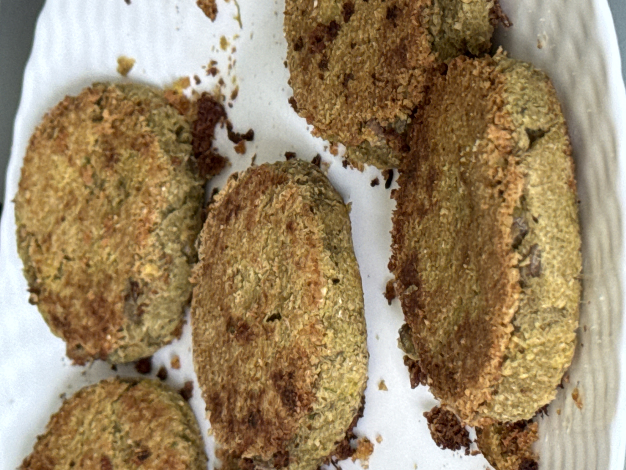 An overhead, close-up shot captures several irregularly shaped, roughly circular to oval food items resting on a white surface, possibly a plate or paper towel. The food items exhibit a greenish-brown interior and are covered with a textured, golden-brown, crispy exterior coating, suggesting they have been fried, baked, or air-fried. Scattered dark brown crumbs are visible on the white surface around and between the pieces. The items vary slightly in size and shape, indicating they may be handmade. They resemble vegetable patties, falafel, or fried cutlets. No activity is explicitly shown, but the food appears to be prepared and ready to be eaten. There is no visible text, and the time of day, weather, and specific location cues within the image are not discernible.