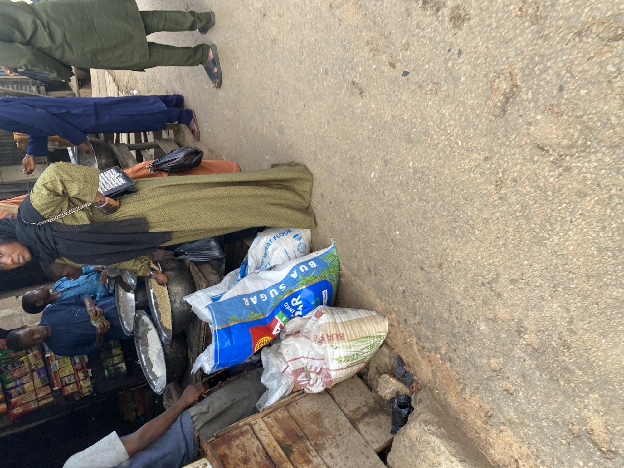 An outdoor market scene in Jos, Nigeria, features several individuals engaged in commerce. In the foreground, large sacks of what appear to be flour and sugar are stacked, one prominently displaying the brand "BUA SUGAR" in bold blue lettering. To the left, a wooden structure, possibly part of a stall, is visible, alongside a pile of rocks.
In the mid-ground, several people are gathered around large metal bowls filled with granular substances, likely food staples. One person, wearing a green patterned dress and a black headscarf, holds a black purse with a chain strap. To their left, a younger individual in a blue shirt is handling money. Another person in a blue robe stands behind the bowls.
In the background, a stall stocked with various packaged goods is visible. The ground is a rough, unpaved surface typical of a market. The overall impression is a bustling marketplace, with transactions and daily activities taking place.