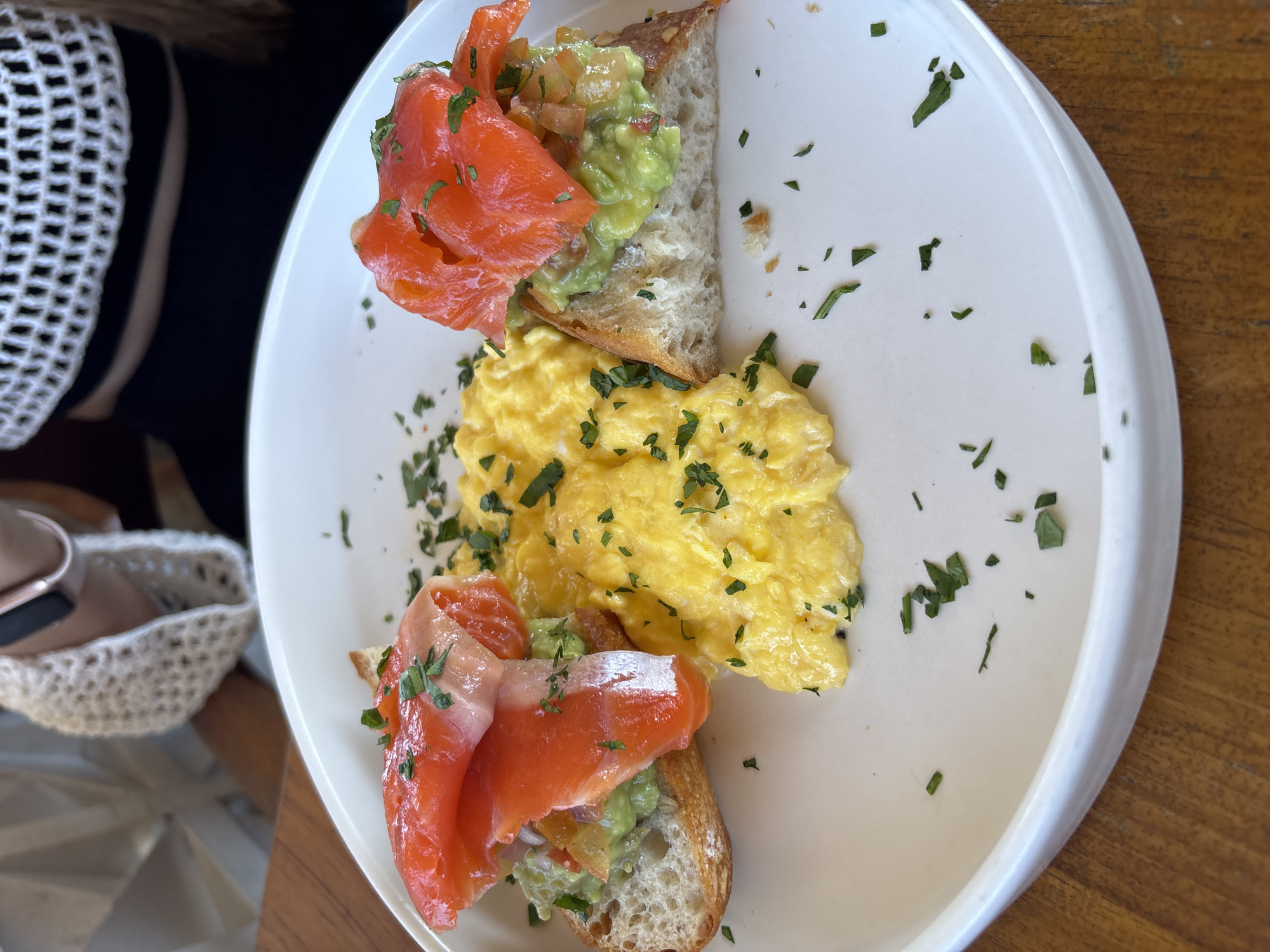 This image captures a close-up of a breakfast or brunch meal served on a white, circular plate, set on a wooden table within a dining establishment in Kuta Selatan, Indonesia. The plate holds two pieces of toasted bread, each topped with a layer of mashed avocado, which appears to contain finely diced red and possibly purple ingredients. Draped over the avocado on each toast are multiple slices of vibrant orange-red smoked salmon. A liberal sprinkling of chopped green herbs, likely parsley, garnishes both the salmon and avocado. Positioned centrally on the plate, between the two toast pieces, is a serving of fluffy, bright yellow scrambled eggs, also garnished with chopped green herbs. Toast crumbs and scattered herb fragments are visible on the white plate. In the upper portion of the frame, two individuals are partially visible behind the plate. The person on the left wears a light-colored, possibly white, garment with a textured, open-knit detail over their shoulder/arm, and a dark band, potentially a smartwatch, on their wrist. The person on the right is wearing a black top or dress, overlaid with a white, open-knit, crocheted crop top, revealing a patterned design. Their dark hair extends over their right shoulder. The setting suggests an indoor or covered outdoor dining area with bright, possibly natural, lighting.
