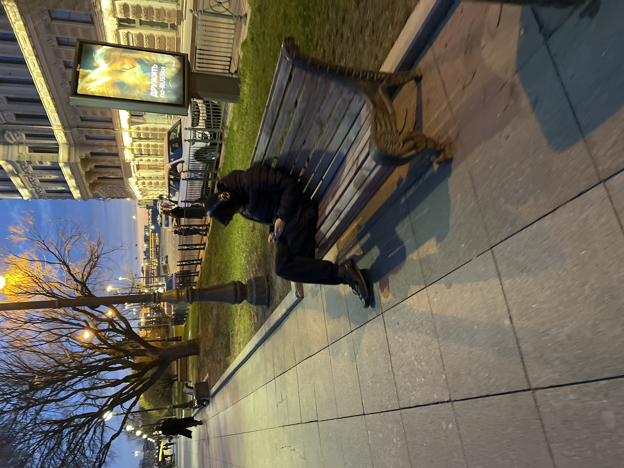The image shows a man lying on a park bench. He is wearing dark clothing and shoes. The bench is made of wood and metal. The background shows a street with buildings, trees, and streetlights. The photograph appears to have been taken in Saint Petersburg, Russia, due to the distinctive architectural style of the buildings and the Russian language on the sign in the background, which translates to "Be friends in Russian."