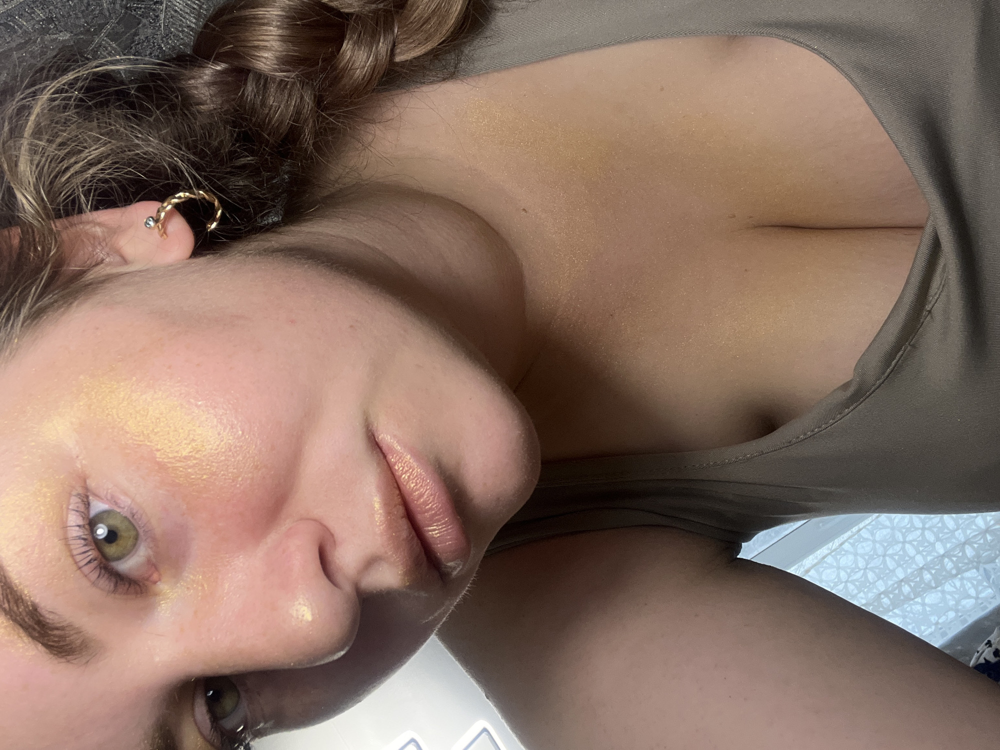 The image presents a close-up, indoor portrait of an individual, likely female, situated in Chaikovsky, Russia. The subject's face is prominently featured, with the left eye, nose, and mouth central to the frame. The left eye displays a light green iris with a dark pupil and dark, defined eyelashes. Both the cheekbones, nose bridge, eyelids, and lips are highlighted with a shimmery, golden-toned cosmetic product. The lips are full and coated with a light peach or nude-colored gloss. Natural skin redness is visible on the cheeks and nose. The individual has brown hair, visible as a braid on the right side of the frame. A gold-colored, braided hoop earring with a small dark gemstone (possibly blue or black) is worn in the visible right earlobe. The subject is wearing a light olive-green or grey-tan ribbed tank top or camisole, with a visible portion of the upper chest and décolletage also showing traces of the golden shimmer. In the background to the left, a blurry white patterned curtain or lace fabric is partially visible, indicating a window. The upper right background is dark and indistinctly textured.