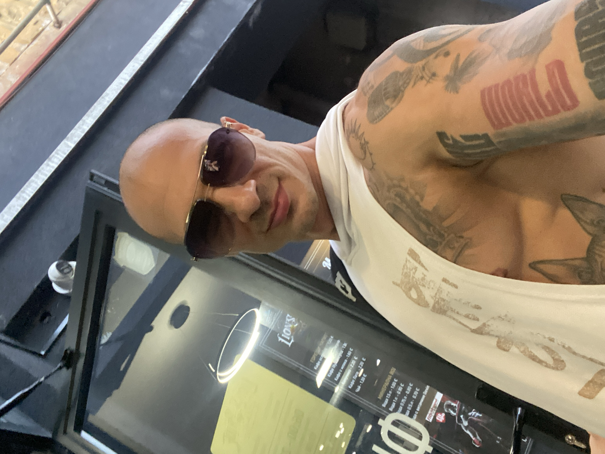 A bald, muscular man wearing sunglasses and a white tank top stands in front of a storefront in Pleven, Bulgaria. The storefront has a large window displaying a menu with prices in Bulgarian currency. A security camera is visible above the window. The man has multiple tattoos on his arms and chest. The lighting suggests it is daytime, and the overall environment appears to be urban.