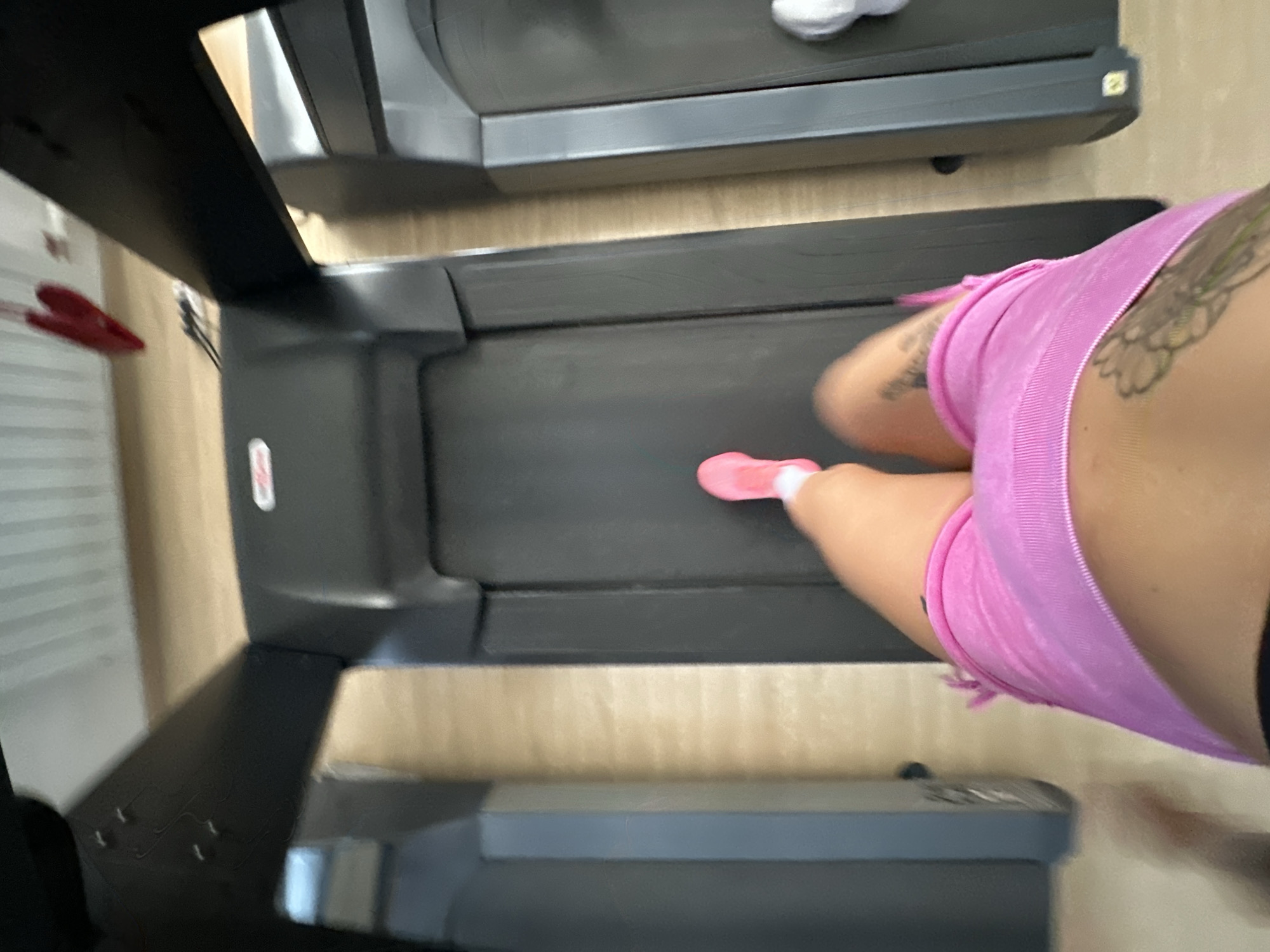 The image shows a person's legs and torso as they run on a treadmill. The person is wearing pink athletic shorts and pink running shoes. There are other treadmills visible in the background. The scene appears to be indoors, likely in a gym. The image is blurry due to motion. The location in Yuzhnouralsk, Russia is not discernible from the image itself.