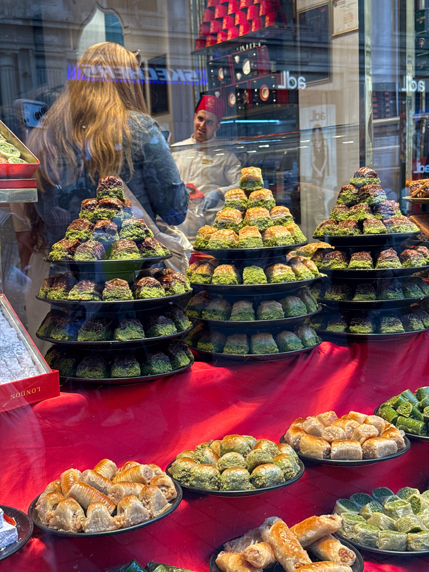 A street-level view of a baklava shop window in Beyoğlu, Turkey, reveals an abundance of sweet treats. The display is dominated by tiered trays of baklava, meticulously arranged in pyramid shapes and rows. The pastries are a mix of golden brown and vibrant green, likely indicating different nut fillings and syrup glazes. Some baklava pieces are coated in finely chopped pistachios, while others appear to be drizzled with chocolate.
In the foreground, a black plate holds a selection of smaller baklava rolls and diamond-shaped pastries, some with visible walnut pieces. Another plate to the right displays more green baklava, and further to the right, a third plate shows more of the diamond-shaped variety. A red box labeled "LONDON" is positioned to the left, containing what appears to be Turkish Delight.
Reflected in the window, a woman with long, blonde hair stands looking into the shop. Behind her, a man wearing a red fez and a white chef's uniform smiles. The reflections also show the interior of the shop, with shelves stocked with more red-boxed sweets and framed items on the wall. The overall environment suggests a bustling commercial street, with the red fabric draped across the display counter adding a warm, inviting tone to the scene.